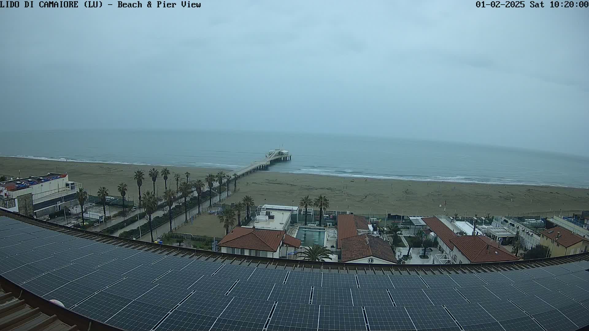 A view from a rooftop with solar panels shows a grey, overcast day at a beach with a pier extending into the ocean, buildings, and palm trees.