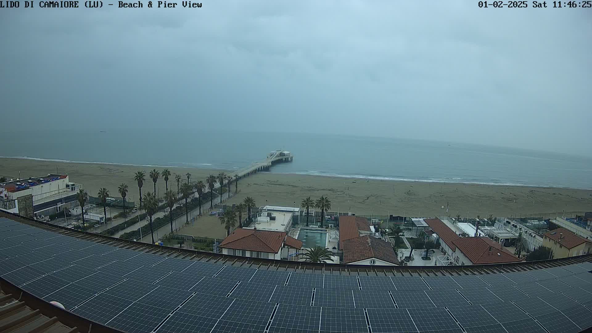 A view from a rooftop with solar panels shows a beach with a pier, buildings, palm trees, and an overcast sky.