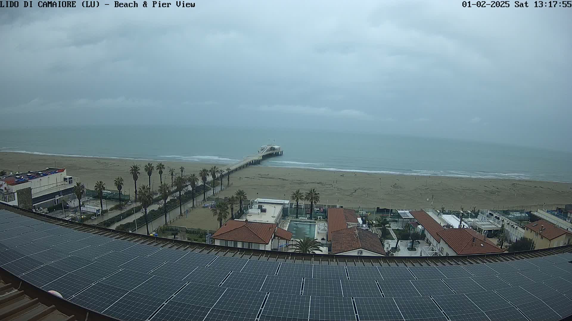 A view from a rooftop covered in solar panels shows a beach with a pier extending into a calm, overcast ocean, palm trees lining the shore, and buildings nearby.
