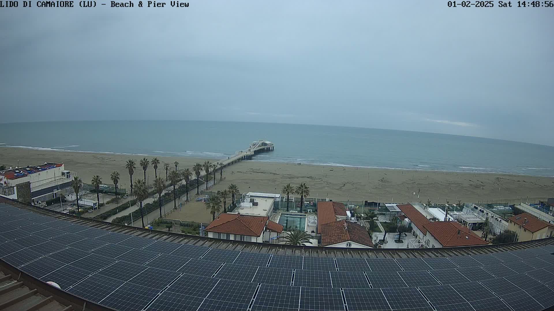 A beach with a pier, buildings, palm trees, and a large solar panel array on a roof are visible under an overcast sky.