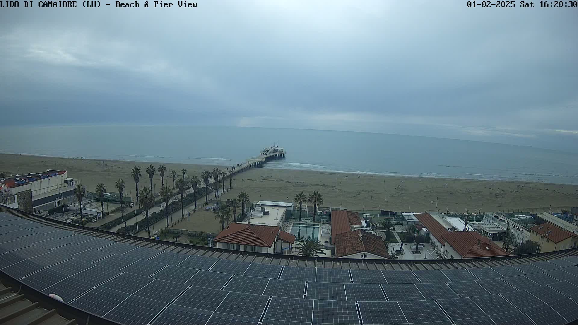 A beach with a pier is visible from a rooftop covered in solar panels under an overcast sky.