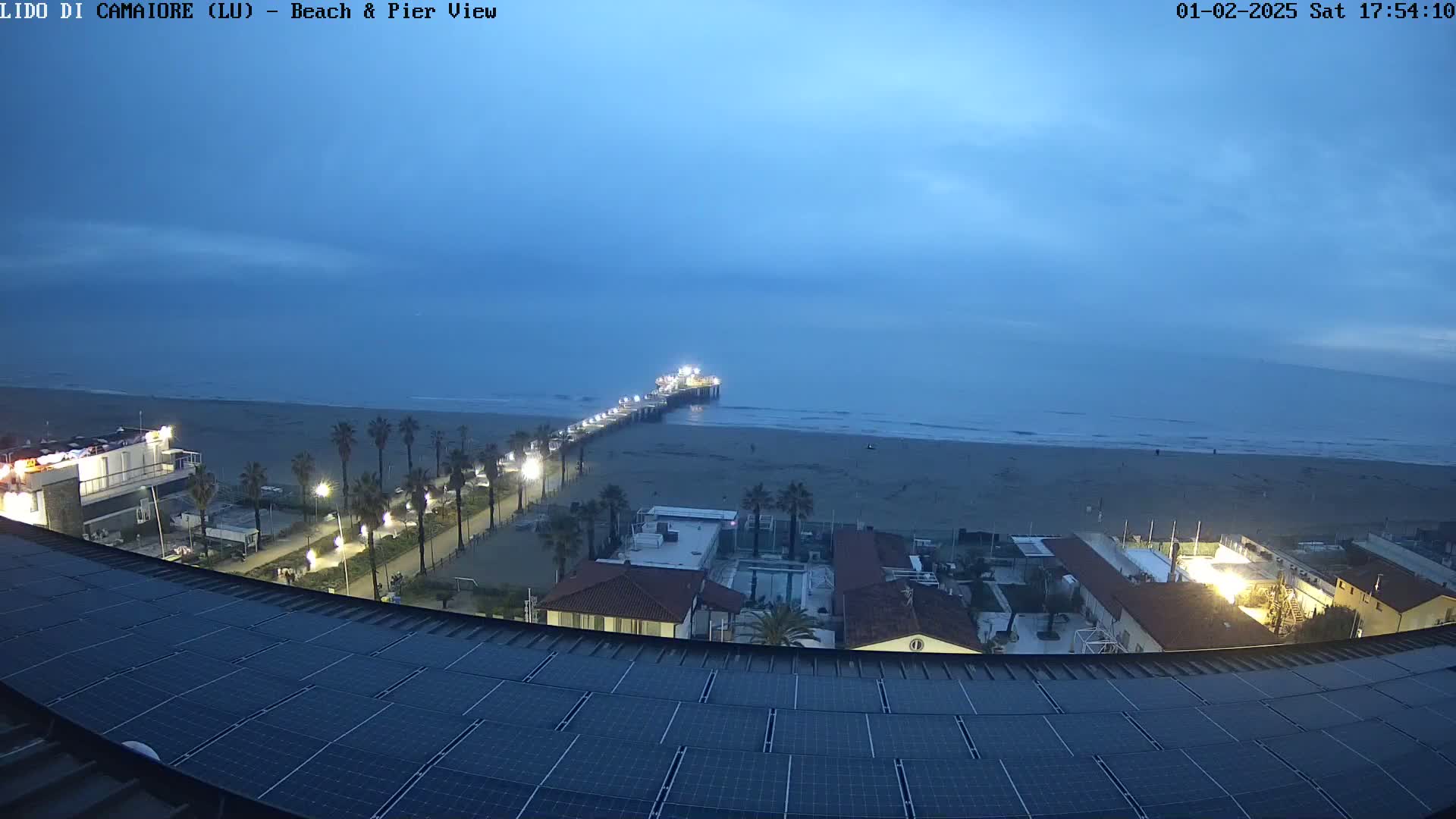 A beach at twilight with a pier extending into the ocean, palm trees lining the shore, and buildings and solar panels in the foreground under a mostly clear sky.