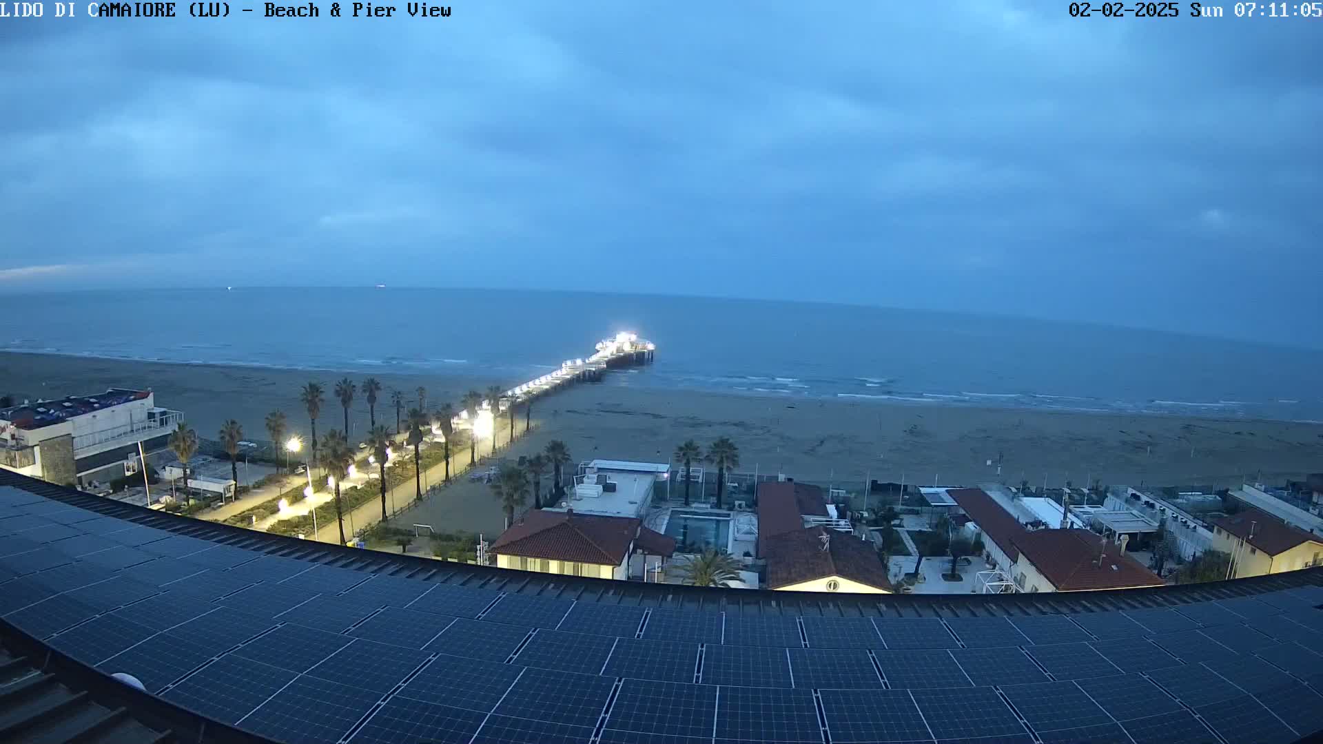 A beach at twilight, with a pier extending into the calm ocean, is viewed from a rooftop with solar panels, showing buildings and palm trees nearby.