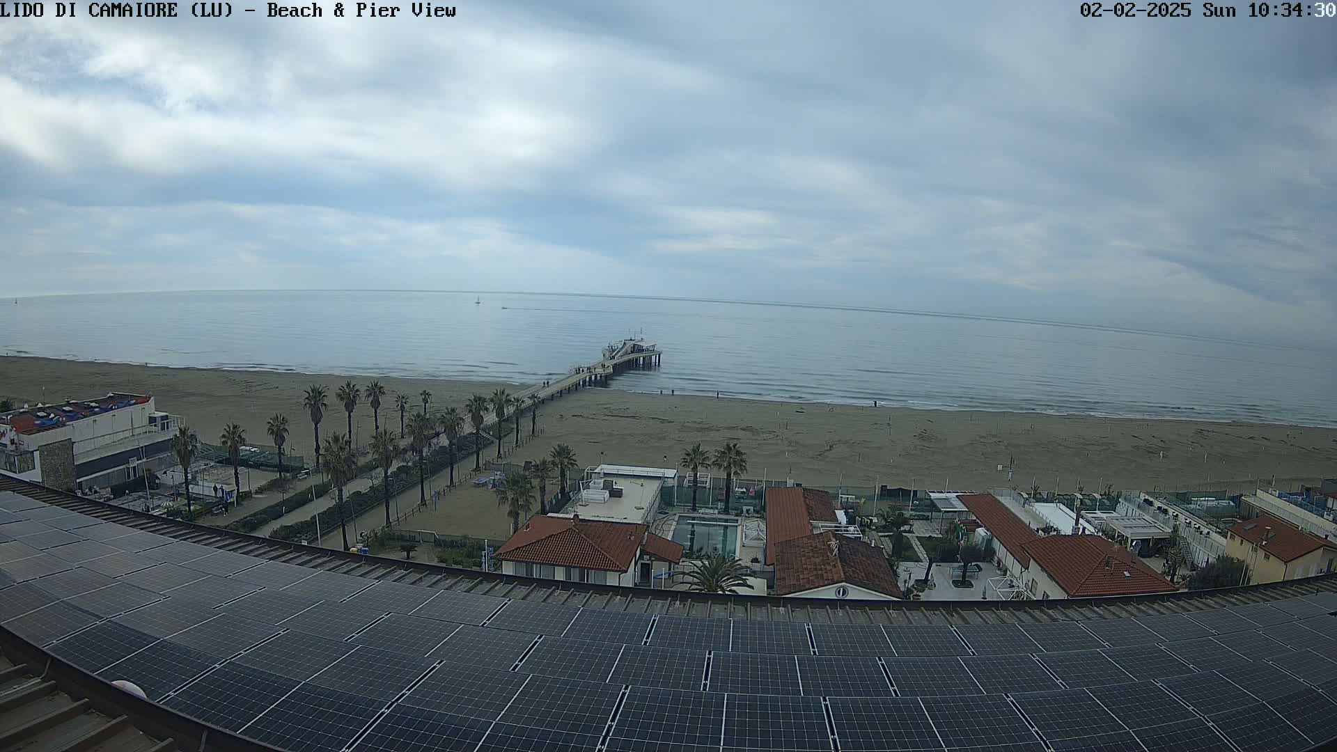 A mostly cloudy day reveals a beach scene with a pier extending into a calm sea, viewed from a rooftop covered in solar panels.