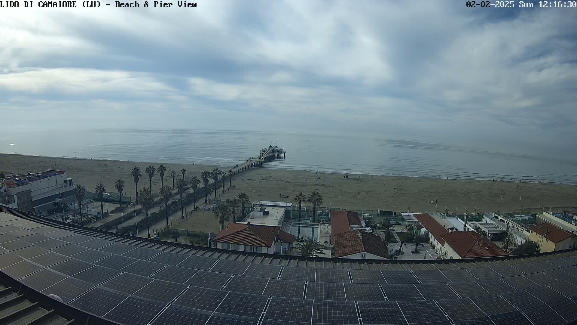 A beach with a pier extends into a calm ocean under a partly cloudy sky, viewed from above a building with solar panels on its roof.