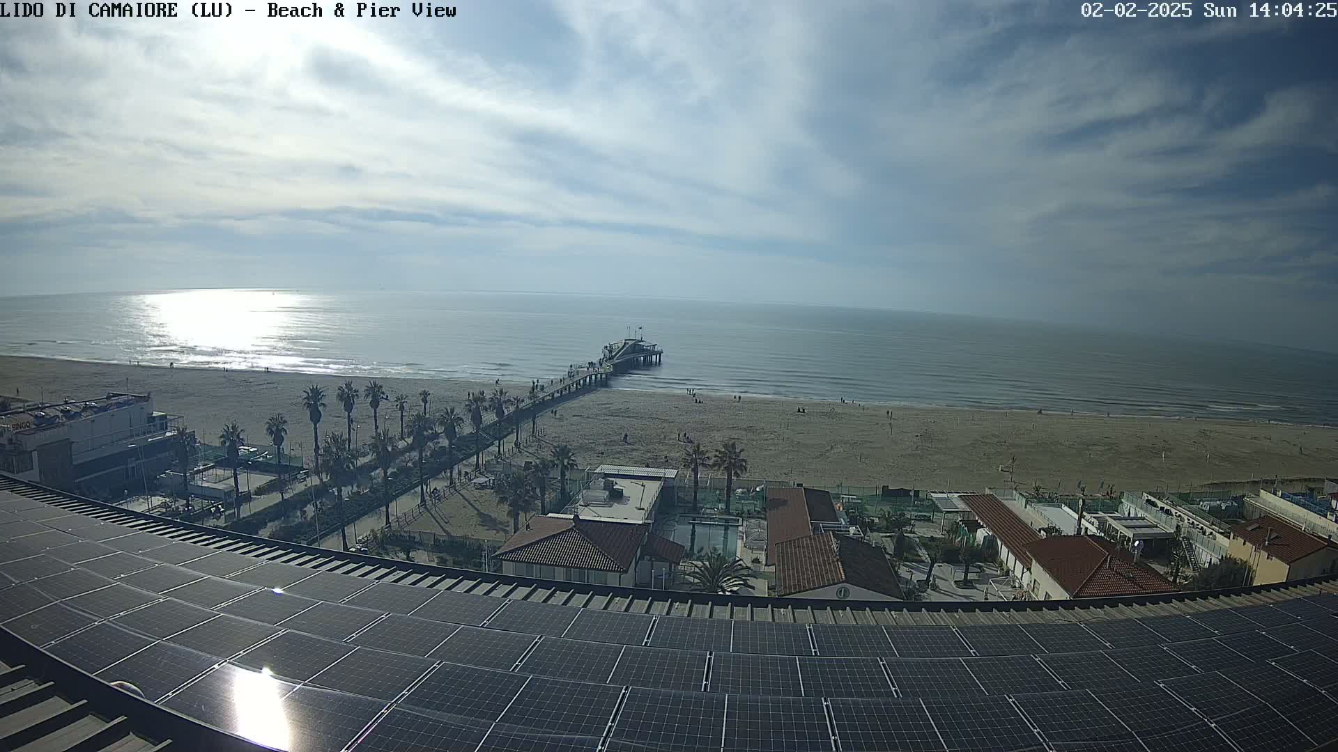 A sunny day at a beach shows a pier extending into a calm sea, with a sandy shore, palm trees, buildings, and a large solar panel array in the foreground.