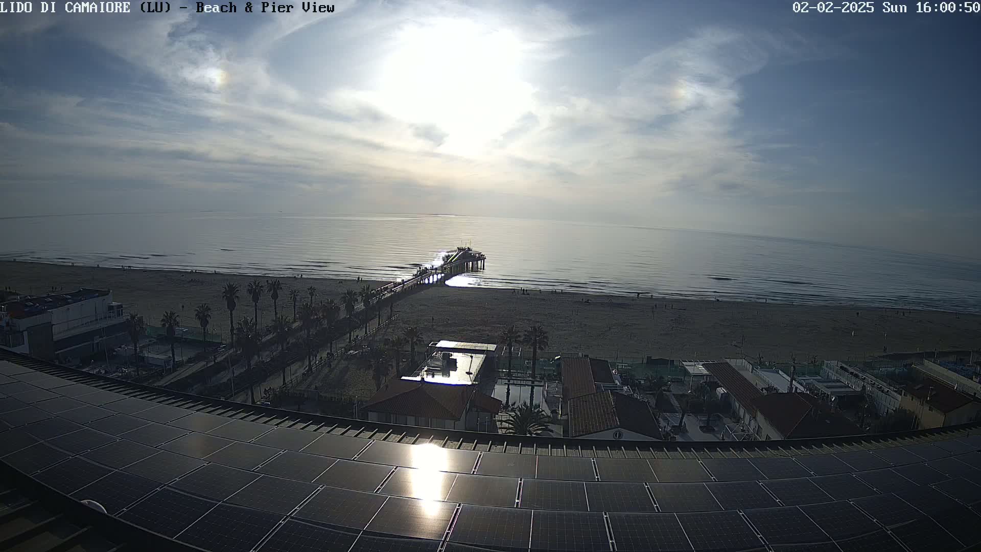 A mostly sunny day reveals a beach scene with a pier extending into a calm ocean, viewed from a rooftop covered in solar panels.