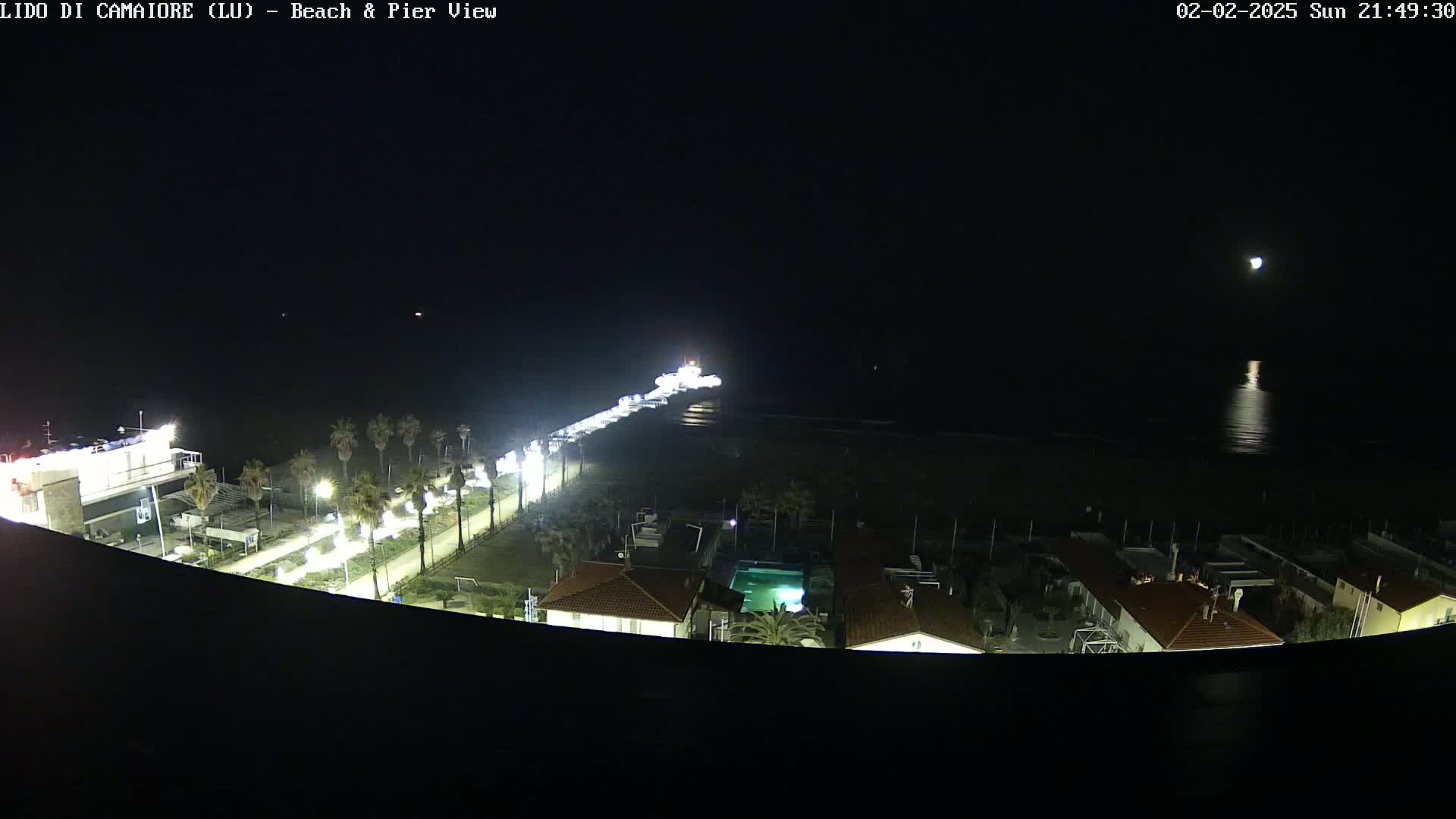 A nighttime aerial view of a beach and pier, with a brightly lit promenade, buildings, and a moonlit ocean.