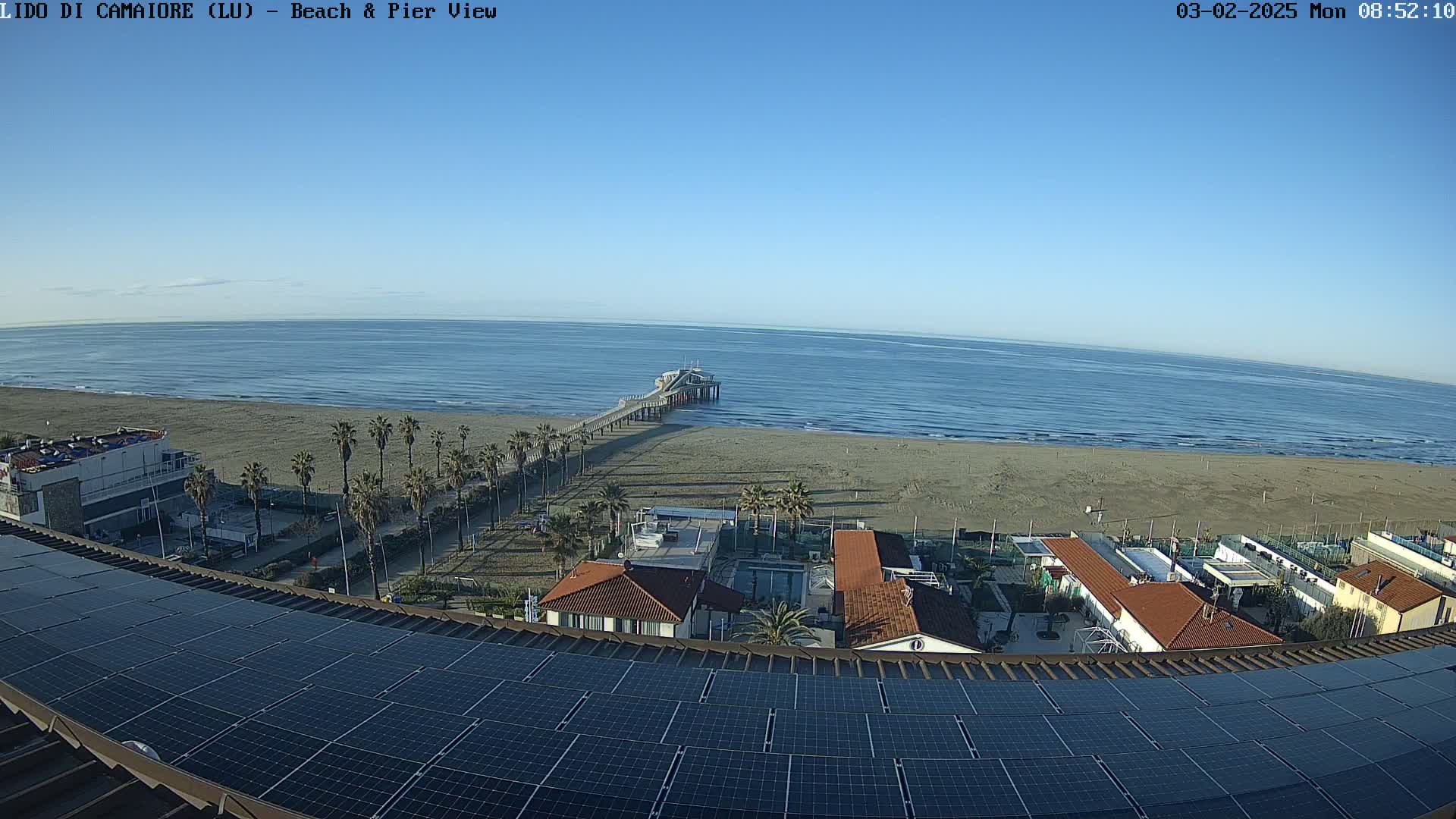 A beach with a pier extends into a calm, blue ocean under a clear sky, viewed from above a rooftop covered in solar panels.