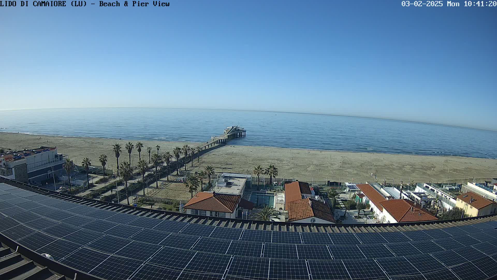 A long pier extends from a sandy beach into a calm, blue ocean under a clear, sunny sky, viewed from a rooftop covered in solar panels.
