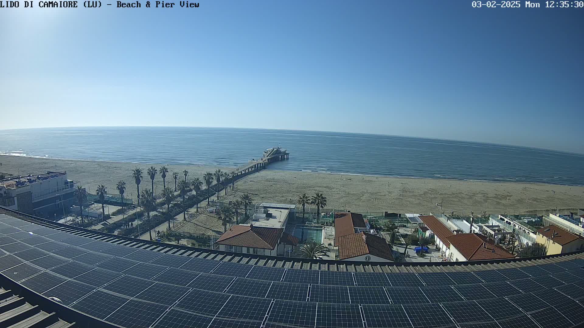 A sunny day overlooking a sandy beach, pier, and ocean, viewed from a rooftop covered in solar panels.