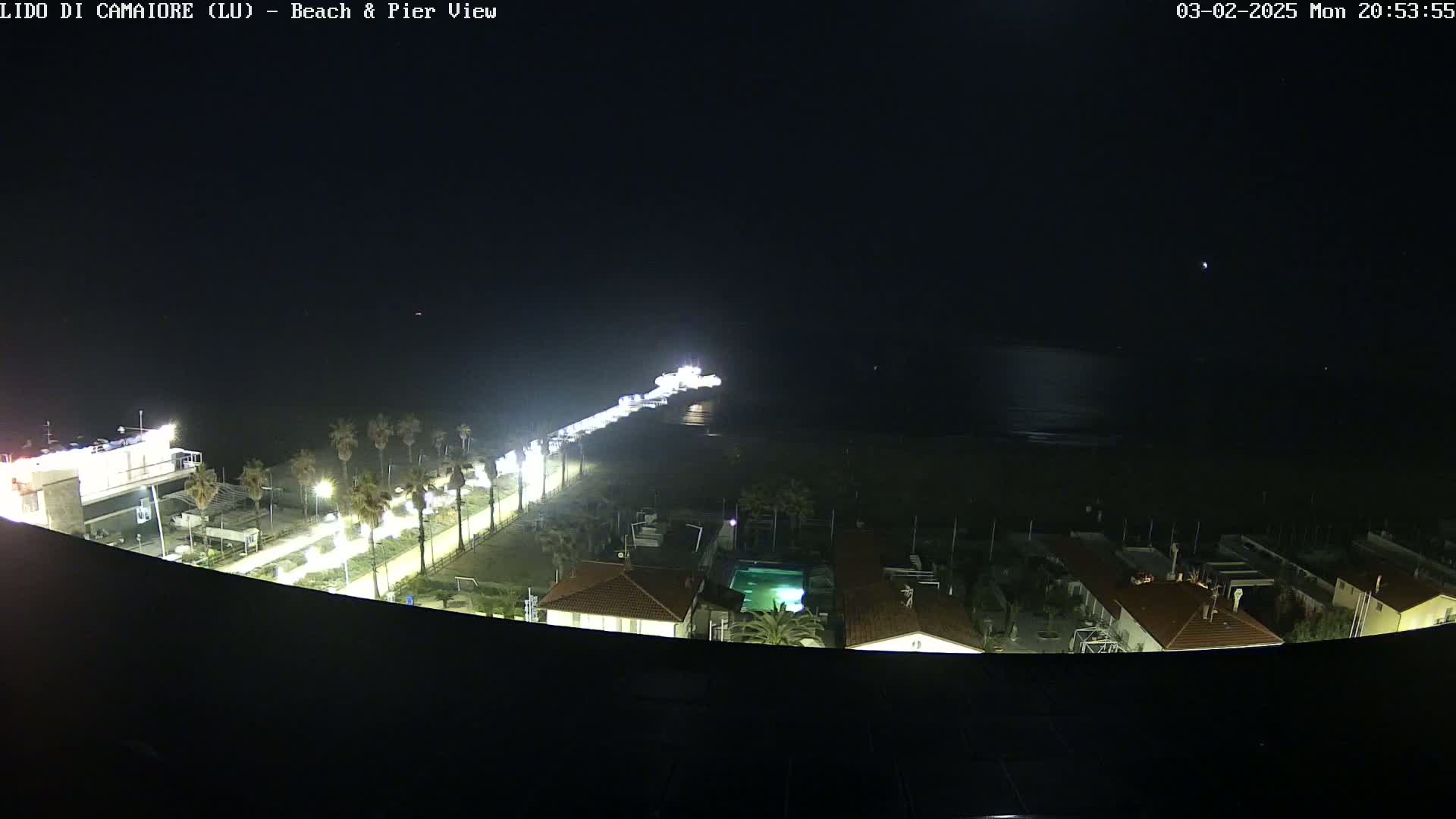 A nighttime aerial view shows a pier extending from a brightly lit beachfront area into a dark ocean, with buildings and palm trees visible along the shore.