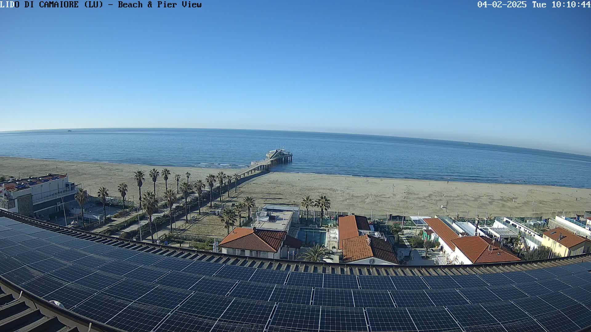 A clear, sunny day overlooking a beach with a pier, palm trees, buildings, and a solar panel array on a rooftop.