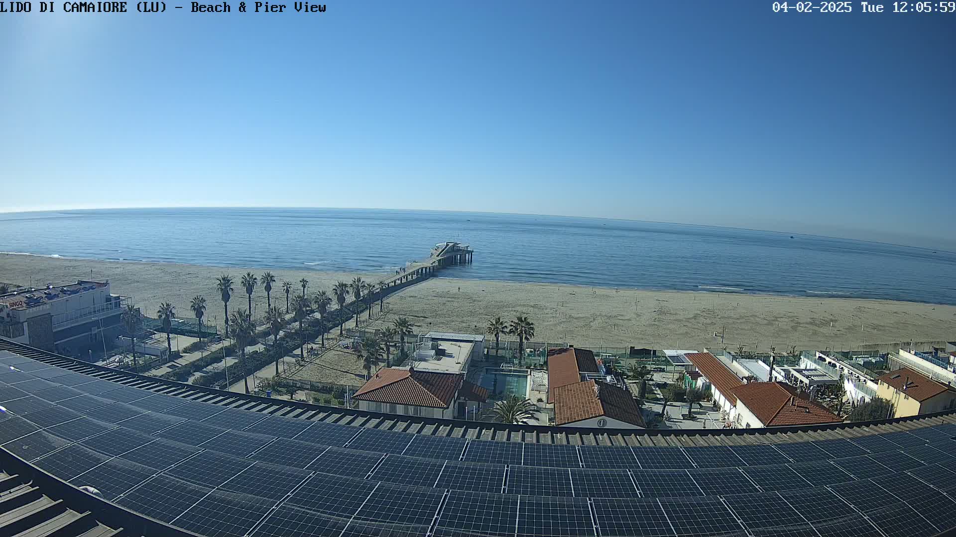 A sunny day reveals a sandy beach, a pier extending into a calm ocean, and buildings with solar panels on the roof, all viewed from an elevated perspective.