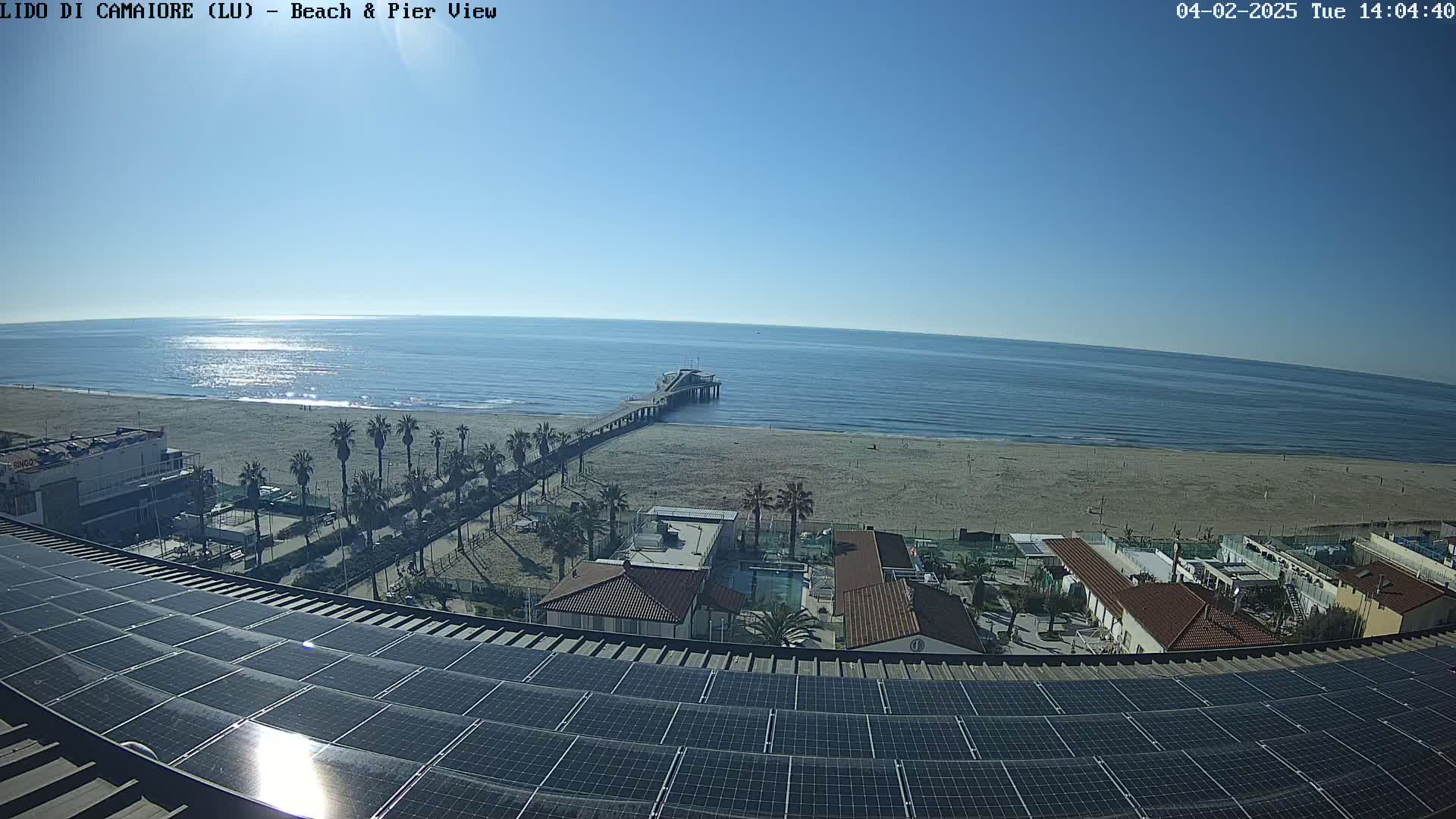 A sunny day shows a beach with a pier extending into a calm ocean, viewed from a rooftop covered in solar panels.