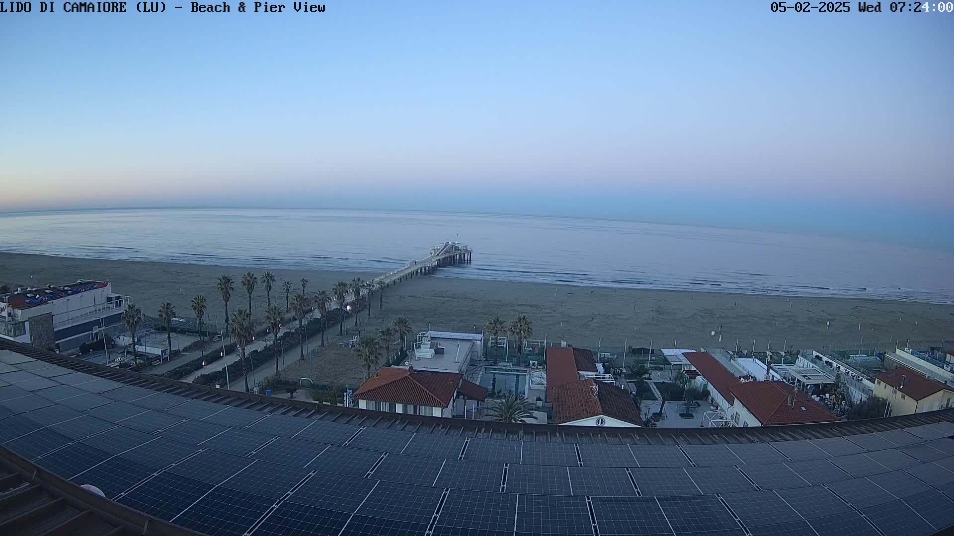 A seaside view at dawn shows a long pier extending from a sandy beach into a calm sea, with buildings and palm trees visible in the foreground, all under a clear, pale blue sky.