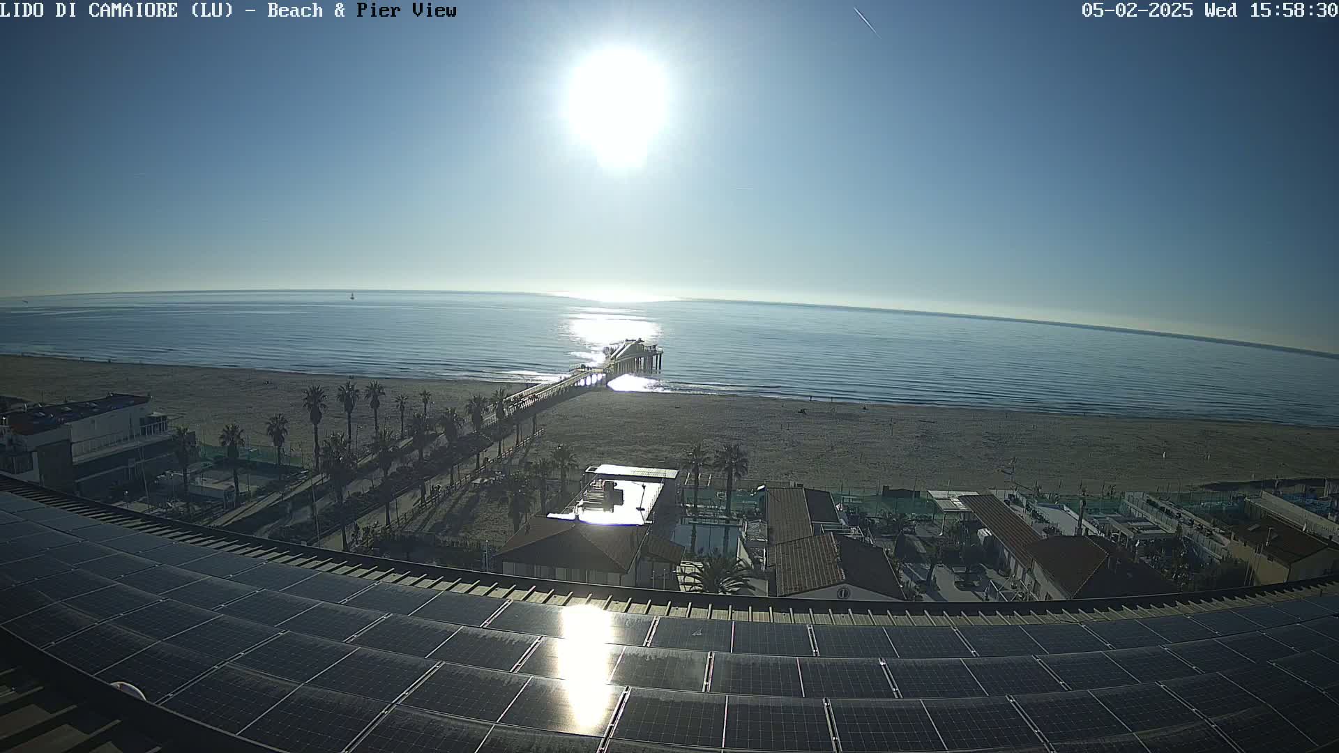 A sunny day overlooking a beach with a pier, palm trees, buildings, and solar panels on a rooftop.
