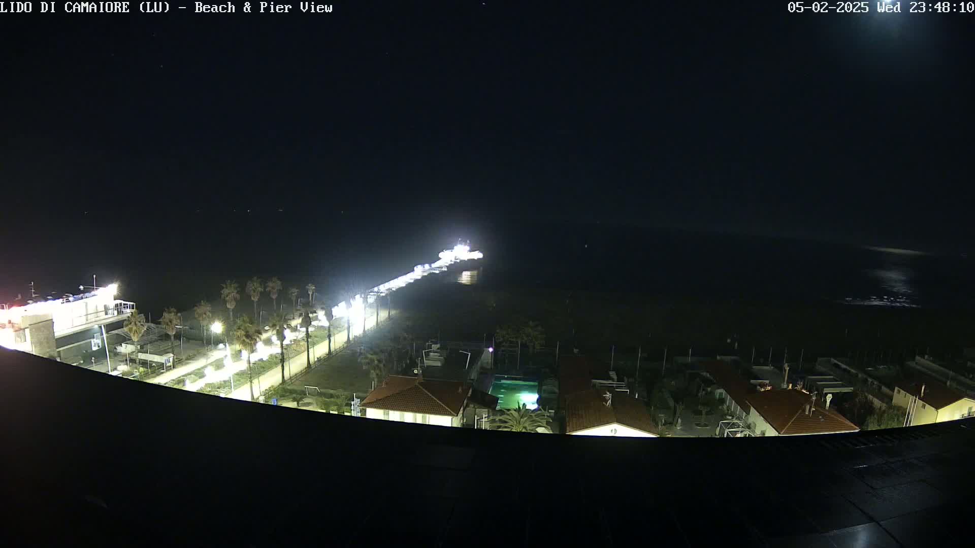 A nighttime, high-angle view shows a beach, pier, and buildings illuminated by lights, with a calm dark ocean in the background.