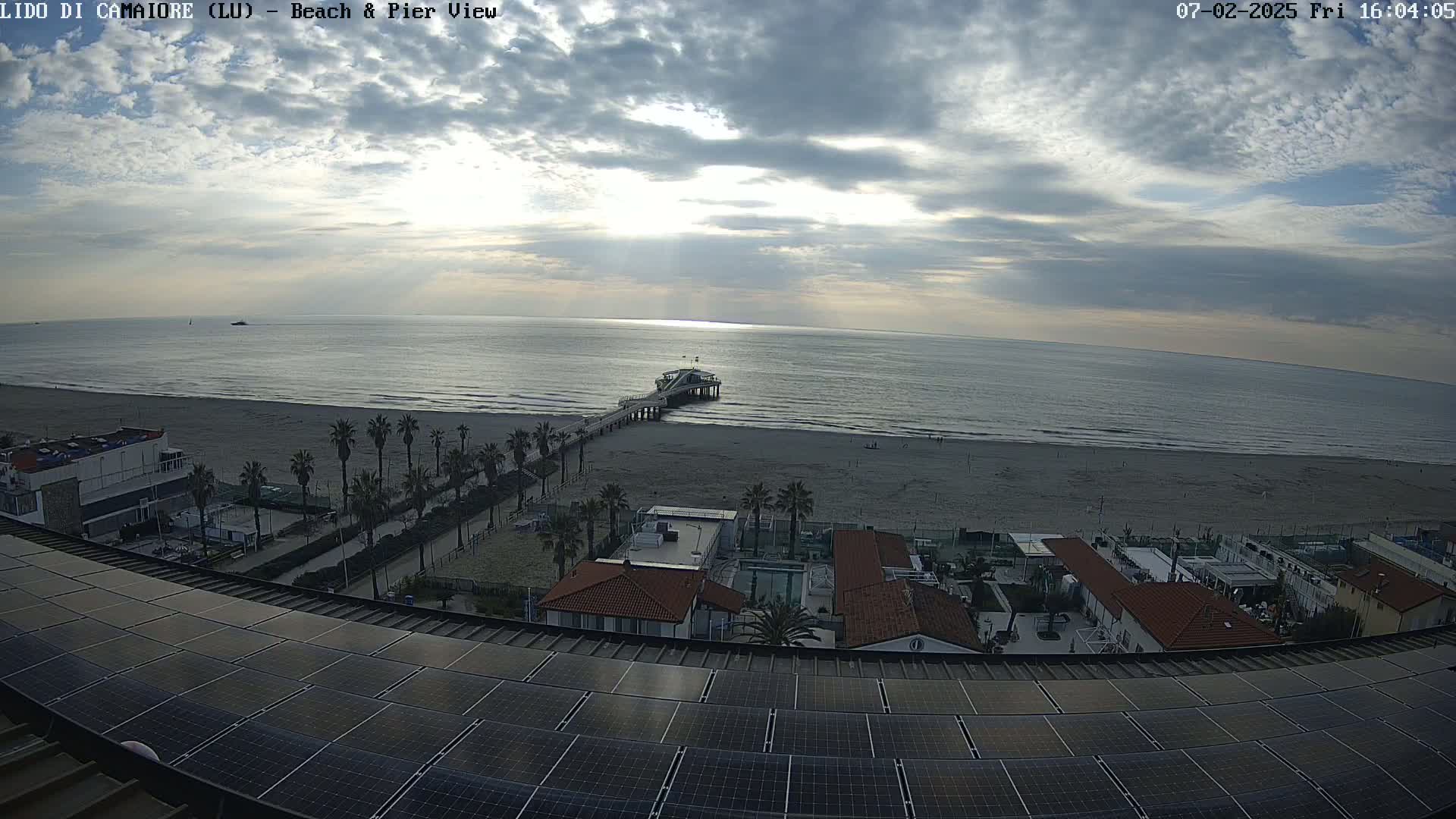 A mostly cloudy sky overlooks a beach with a pier, palm trees, and buildings with solar panels on a roof.