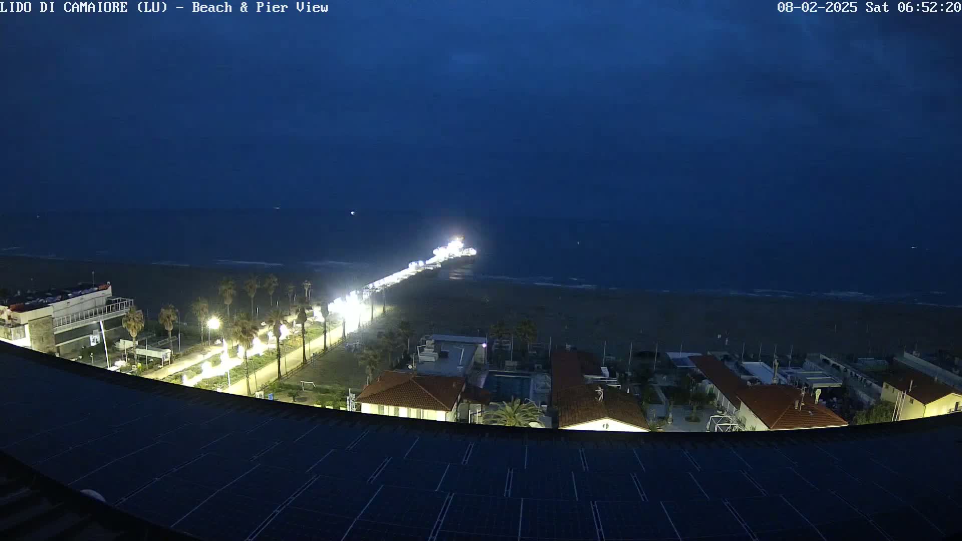 A nighttime, high-angle view shows a pier extending from a beach into a dark ocean under a mostly clear, dusky sky, with buildings and streetlights illuminating the shoreline.