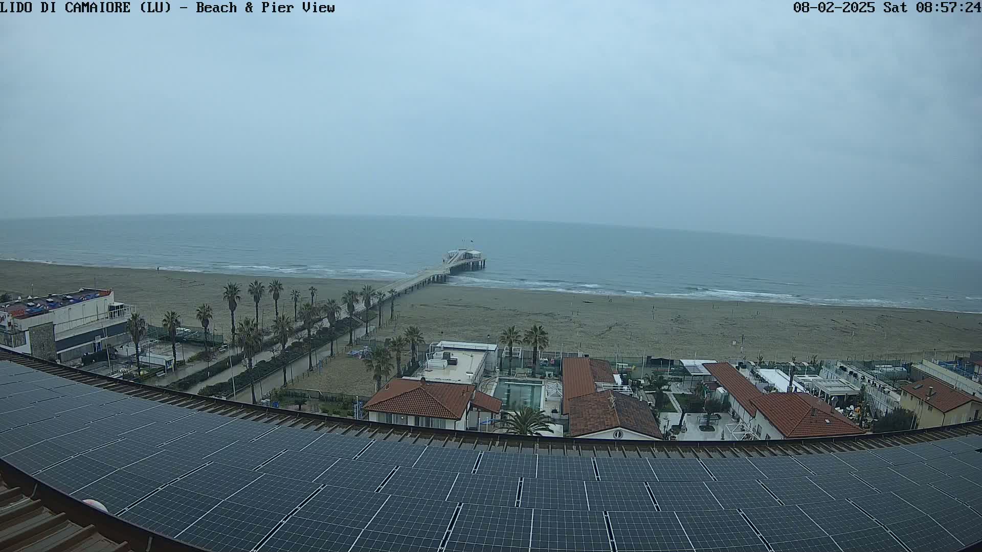 A long sandy beach with a pier extends into a gray, overcast ocean, viewed from above a rooftop covered in solar panels, with buildings and palm trees visible in the foreground.