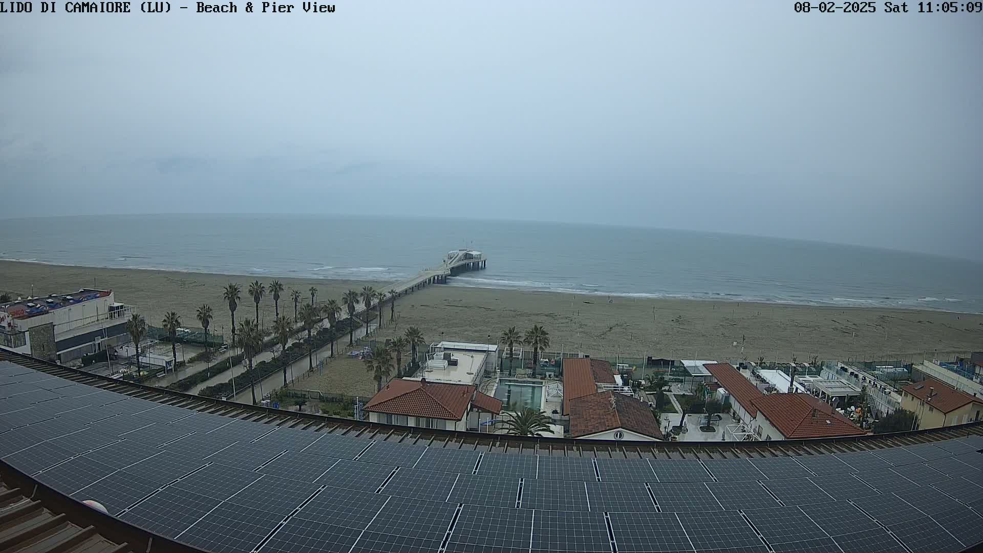 A view from a rooftop with solar panels shows a beach with a pier extending into a calm, gray sea under an overcast sky.