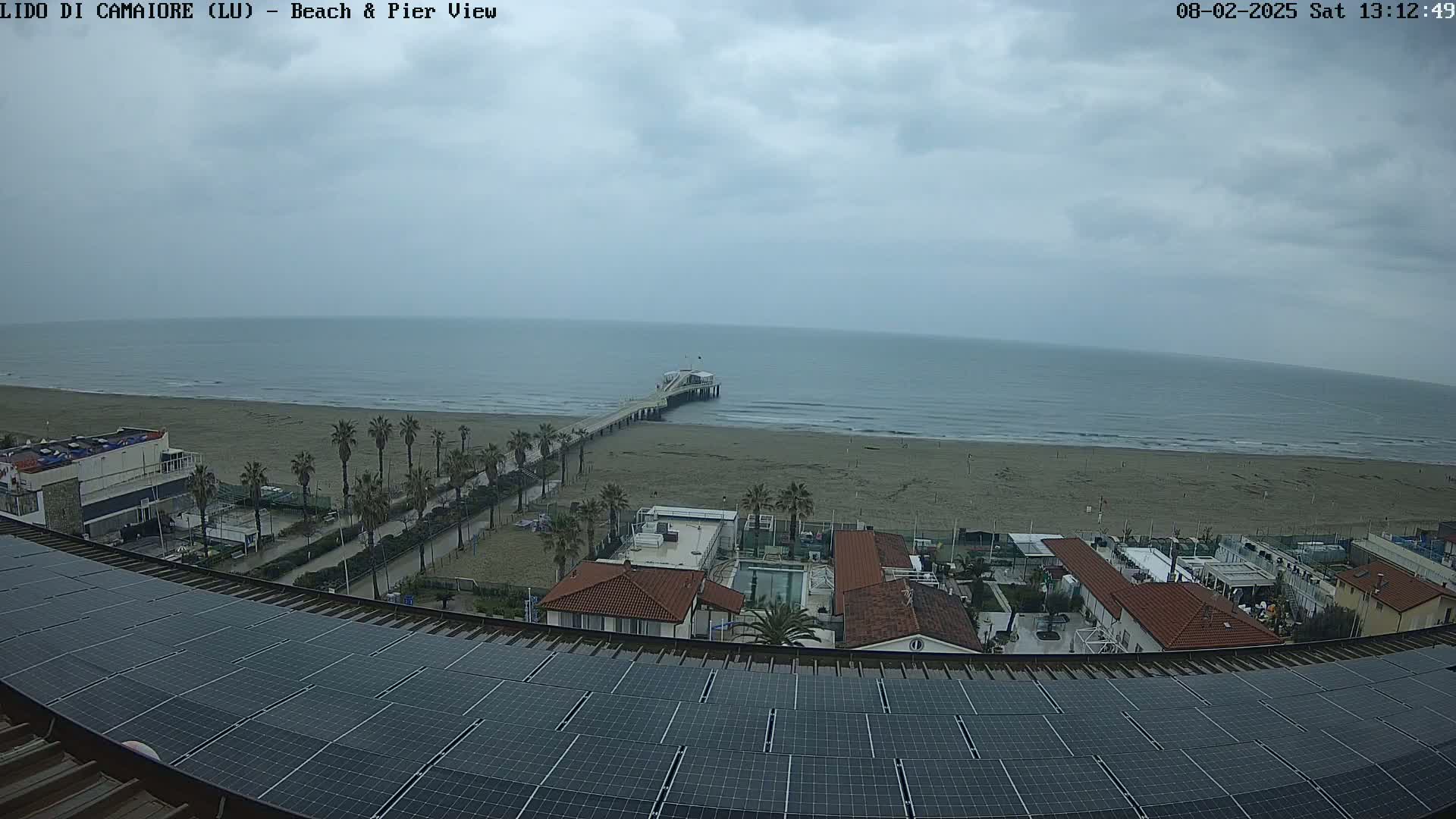 A beach with a pier is visible from a rooftop covered in solar panels under an overcast sky.