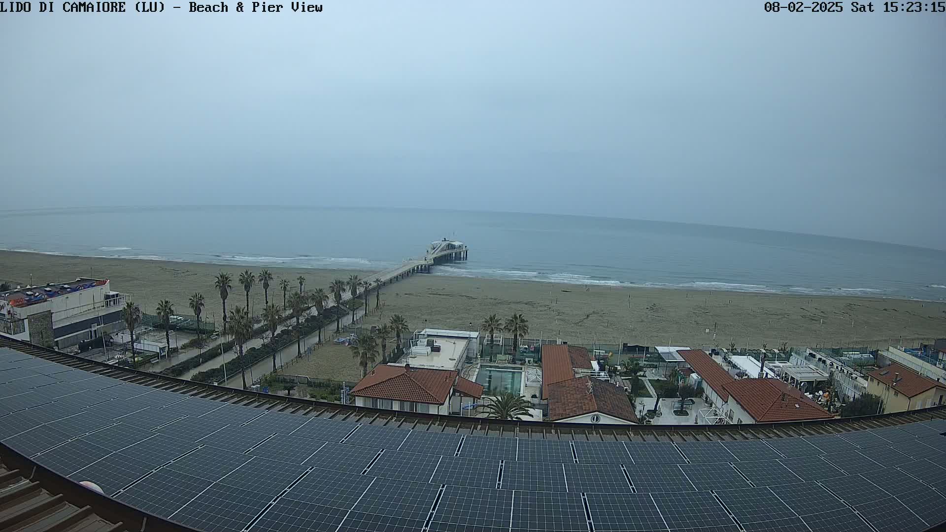 A view from a rooftop with solar panels shows a beach, a pier extending into a calm, overcast ocean, and buildings with palm trees in the foreground.
