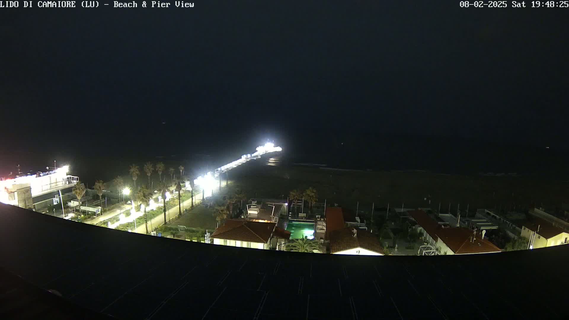 A nighttime view of a beach and pier, with a dark ocean and brightly lit beachfront buildings and pathway.