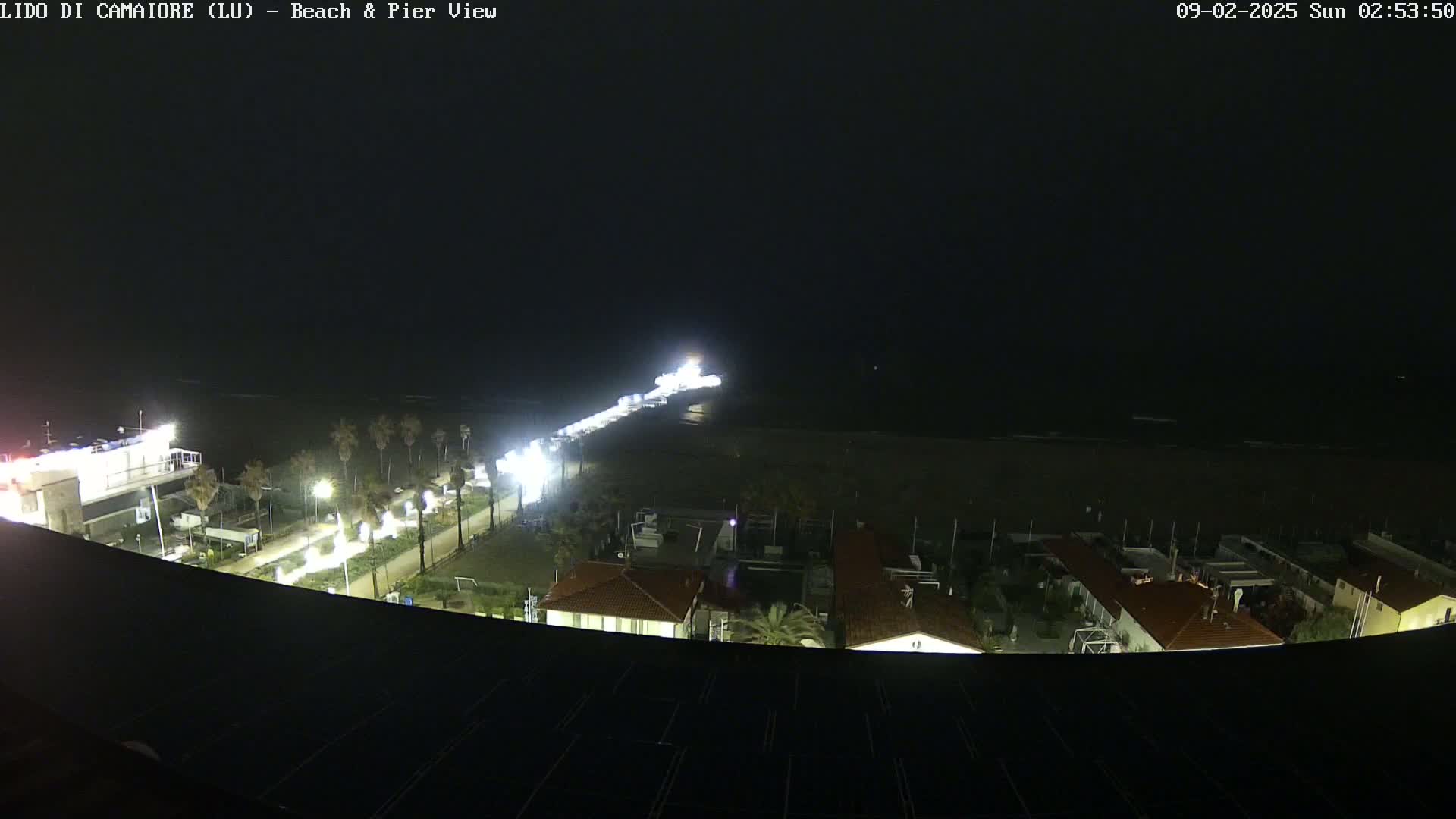 A nighttime, overhead view of a beach and pier, illuminated by lights, under dark skies.