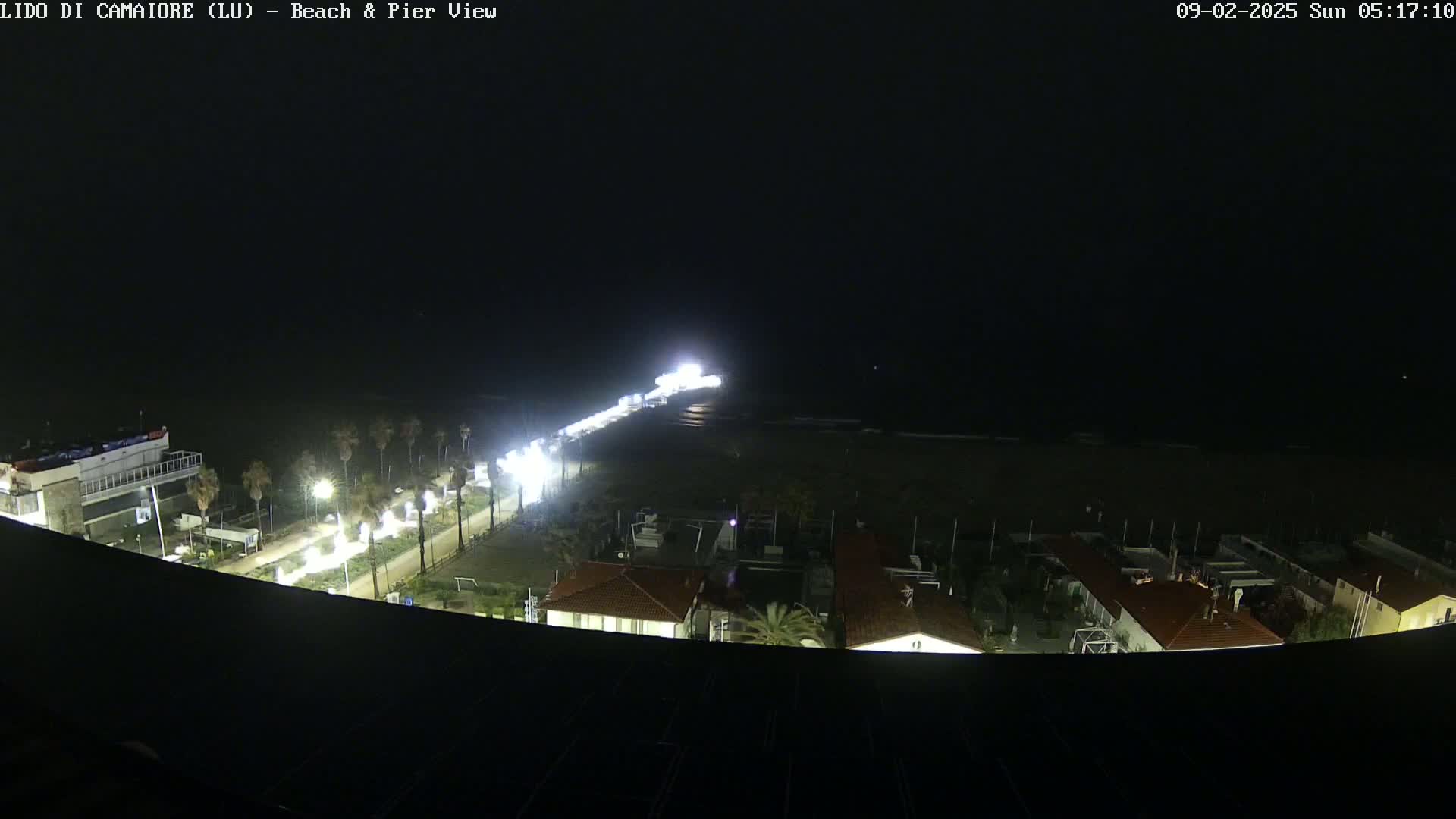 A nighttime, aerial view shows a brightly lit pier extending from a beach, with buildings and palm trees visible along the shoreline.