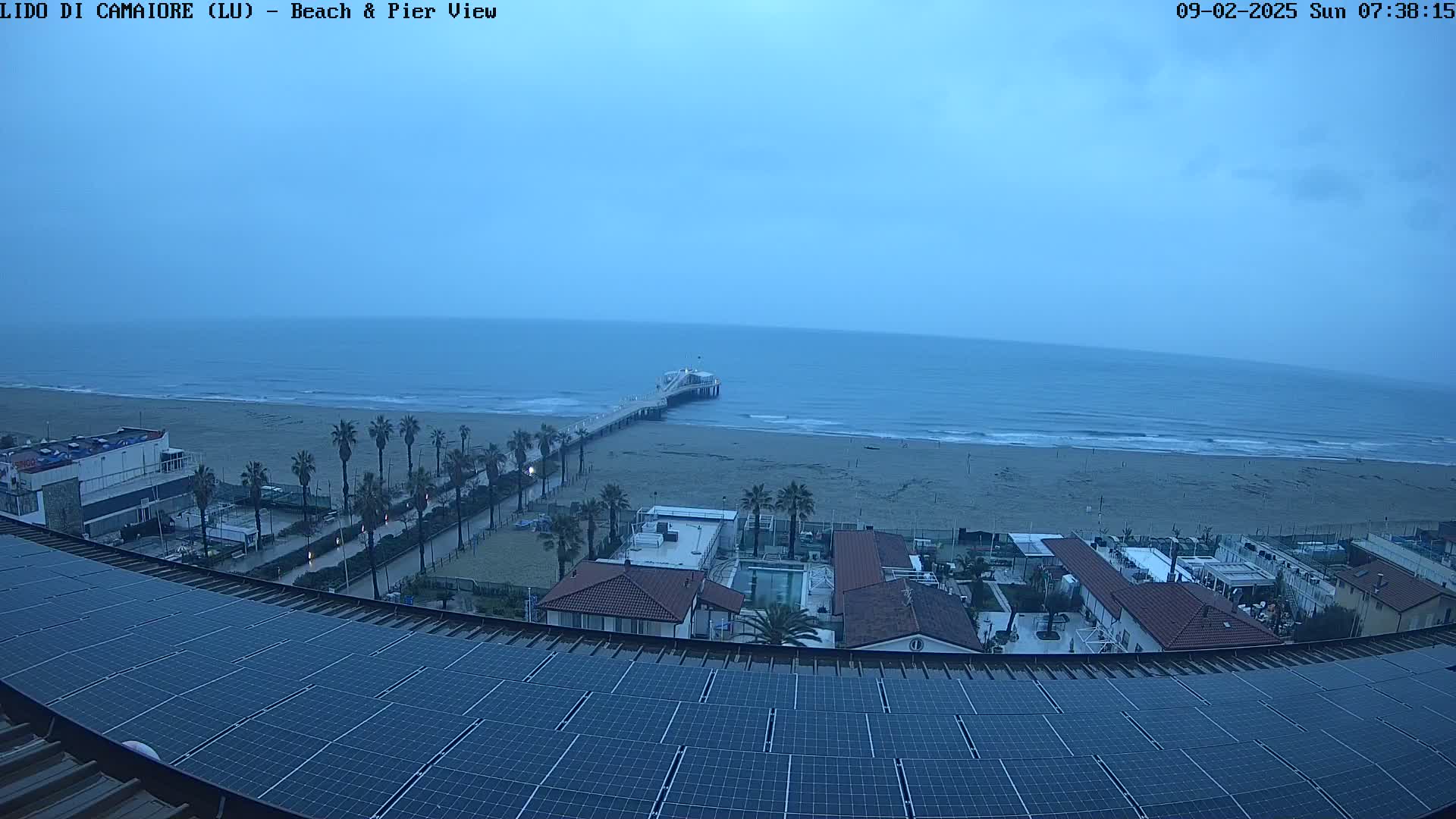 A view from a rooftop with solar panels overlooking a beach, pier, and ocean under an overcast sky.