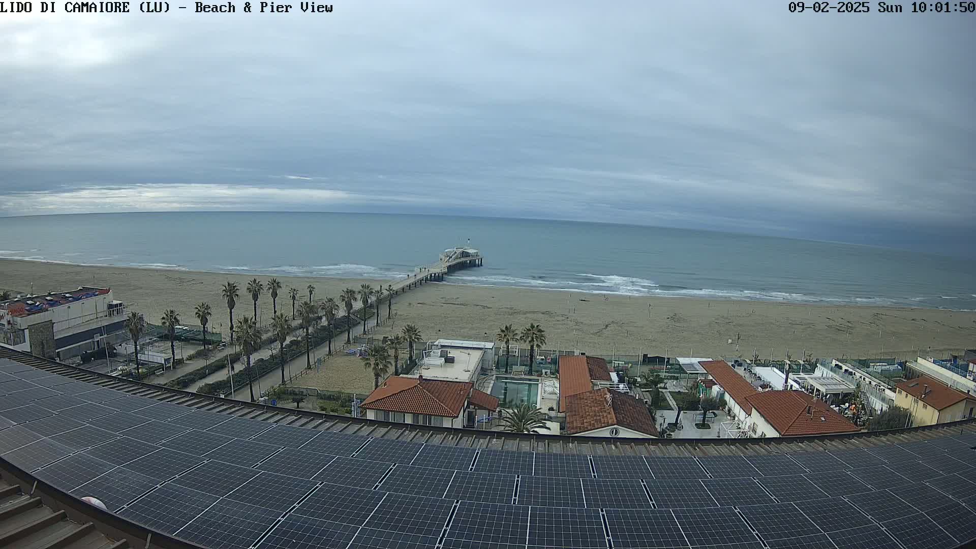 A beach with a pier extends into a calm sea under a cloudy sky, viewed from above a rooftop covered in solar panels.