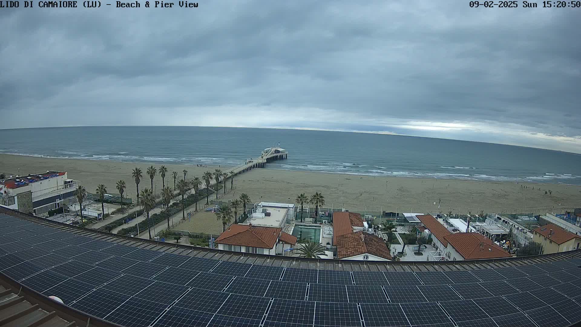 A beach scene under an overcast sky shows a pier extending into a calm sea, adjacent to buildings with red tile roofs and a large solar panel array on a rooftop in the foreground.