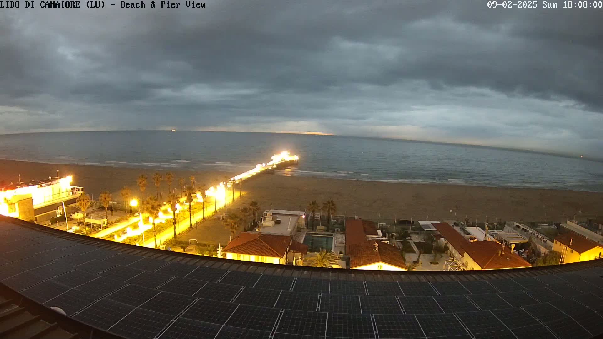 An illuminated beach and pier are visible at twilight under a cloudy sky, viewed from above a building with solar panels on its roof.