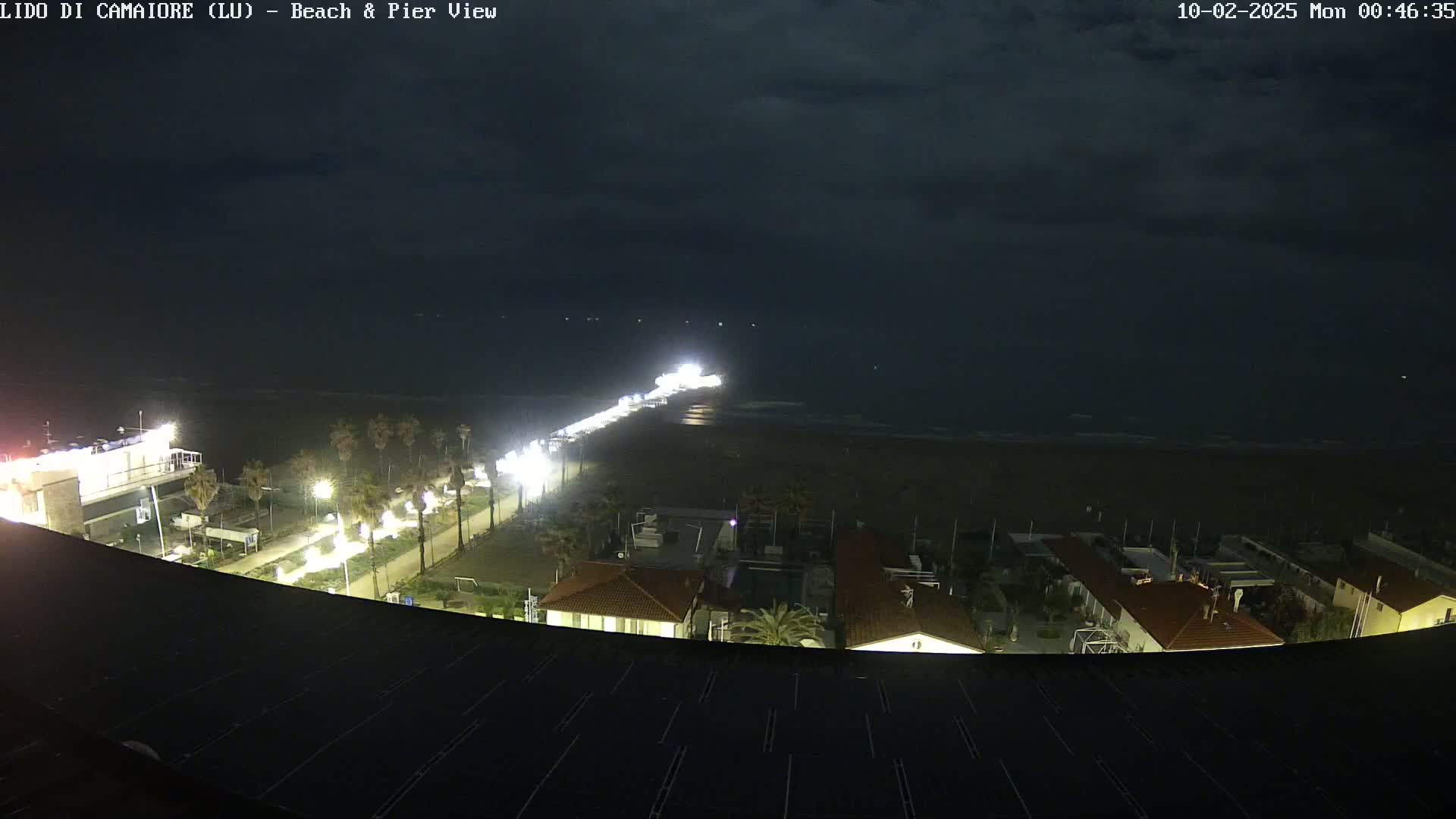 A nighttime view of a beach and pier, with a mostly cloudy sky, shows a long, brightly lit pier extending into a dark ocean, with buildings and palm trees illuminated along the shoreline.