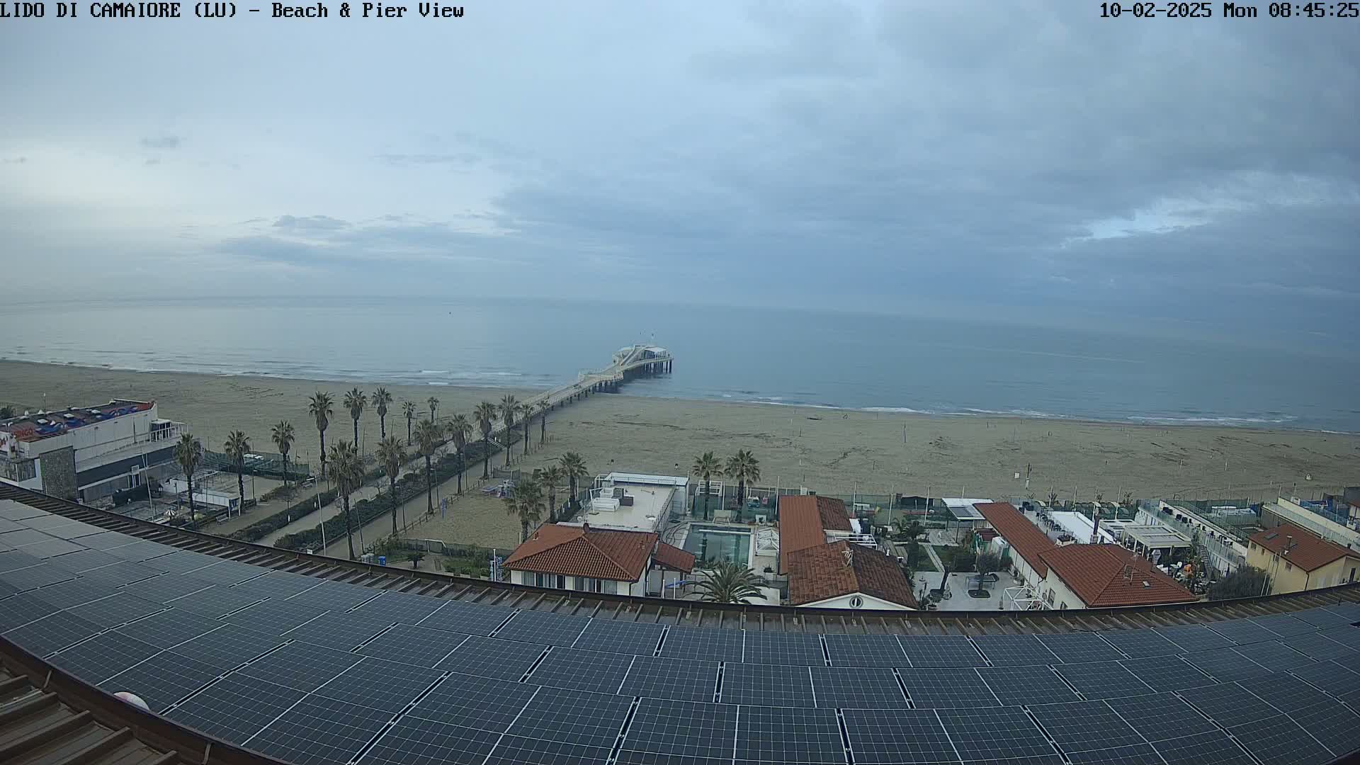 A view from a rooftop with solar panels shows a beach, pier, ocean, and buildings under an overcast sky.
