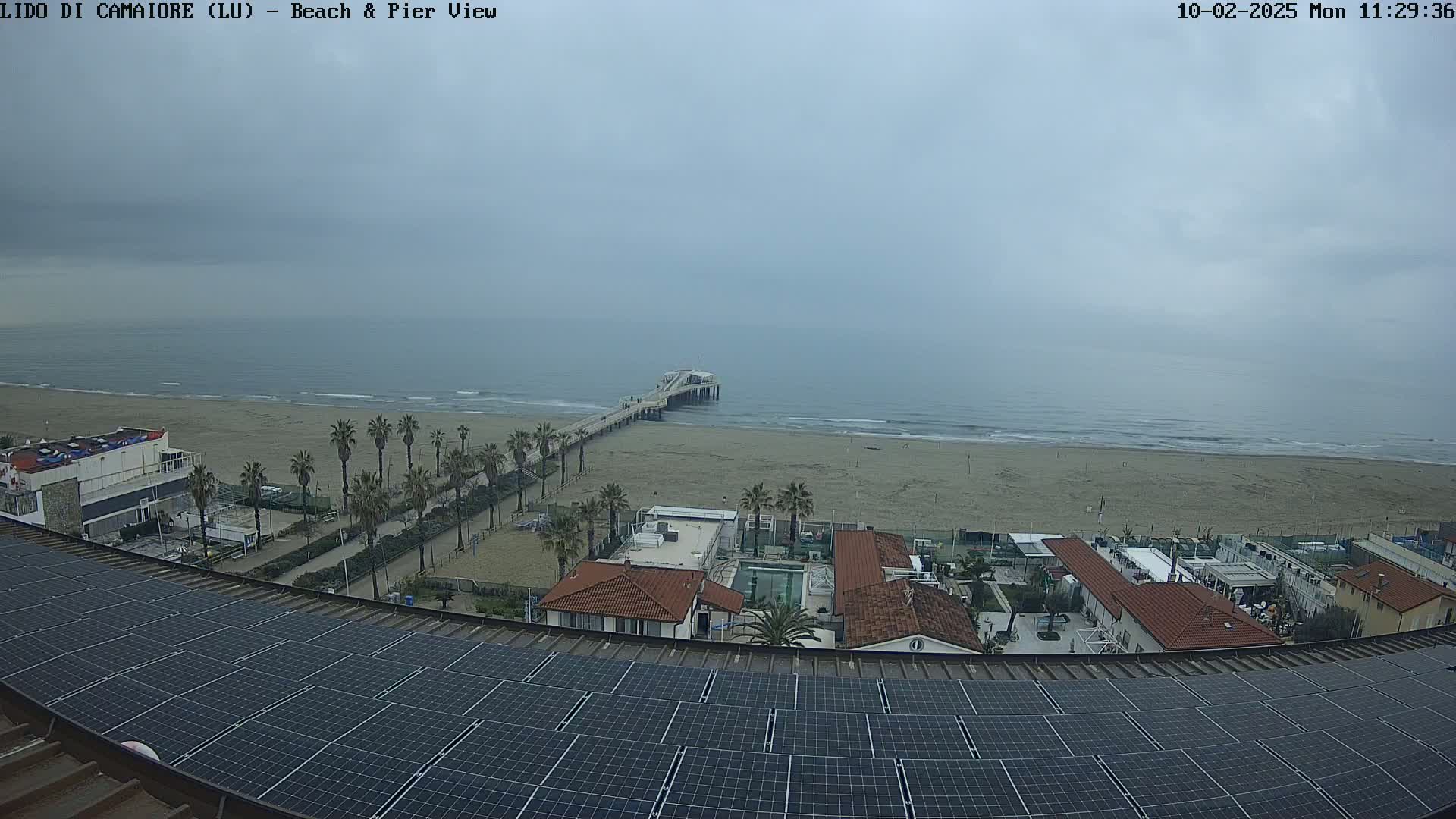 A long sandy beach with a pier extends into a calm, overcast ocean, viewed from a rooftop covered in solar panels, with buildings and palm trees lining the beach.
