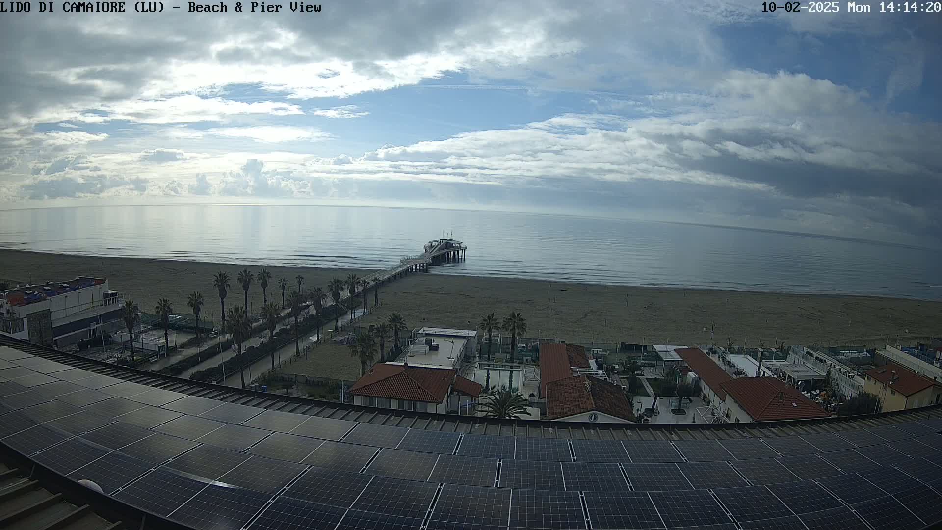 A mostly cloudy day overlooks a beach with a pier, palm trees, and buildings, all viewed from a rooftop covered in solar panels.