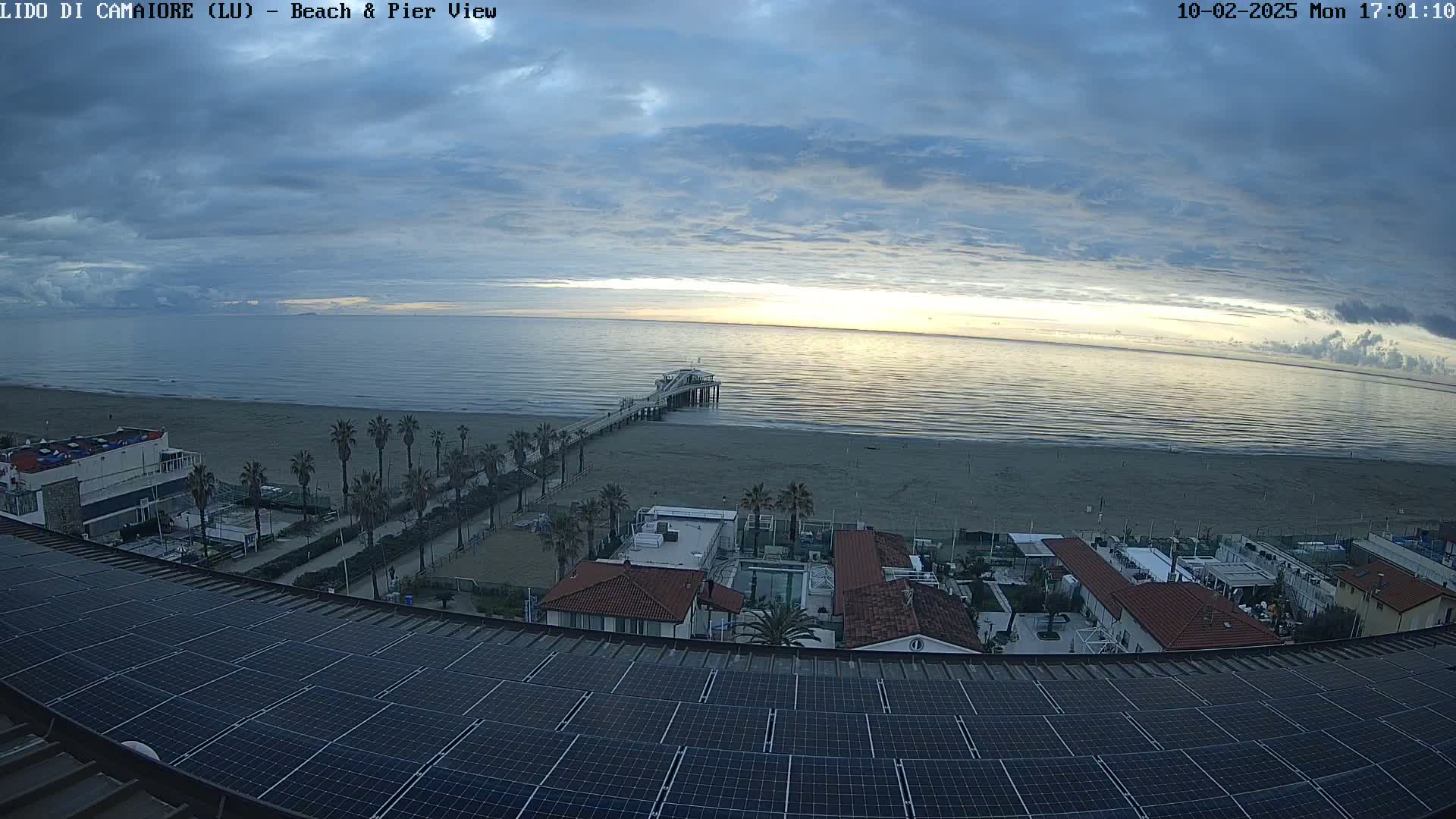 A seaside view shows a pier extending into a calm ocean under a mostly cloudy sky, with a beach, palm trees, and buildings visible in the foreground.