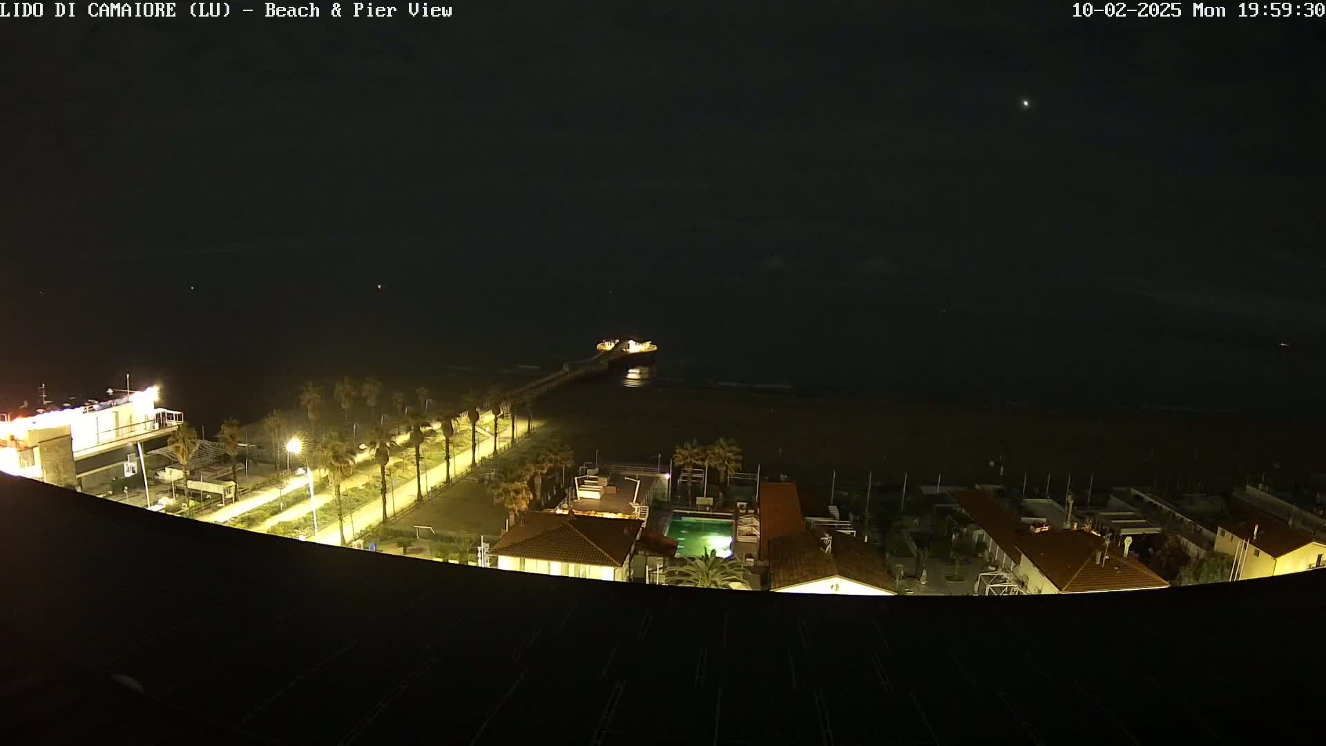 A nighttime, high-angle view shows a pier extending from a beach into a dark ocean, with palm trees and illuminated buildings lining the shoreline.