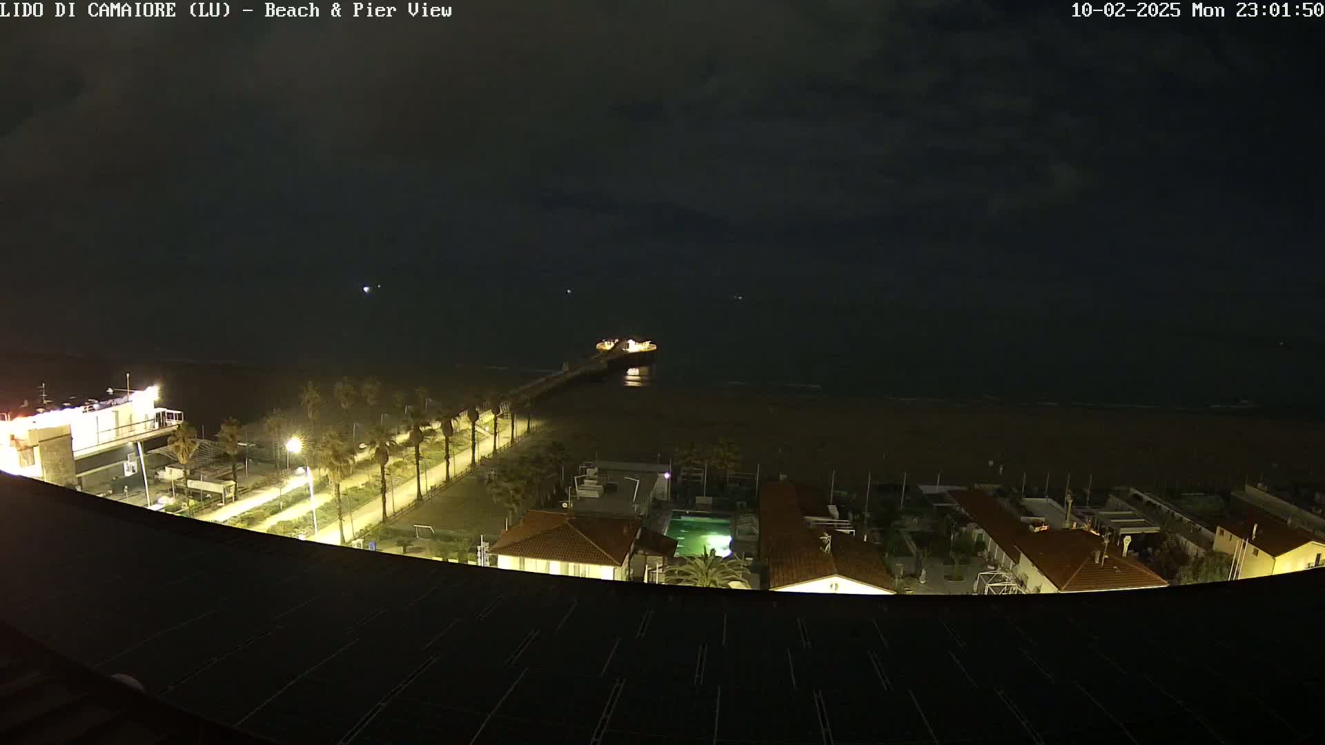 A nighttime view of a beach, pier, and beachfront buildings under a dark, cloudy sky.