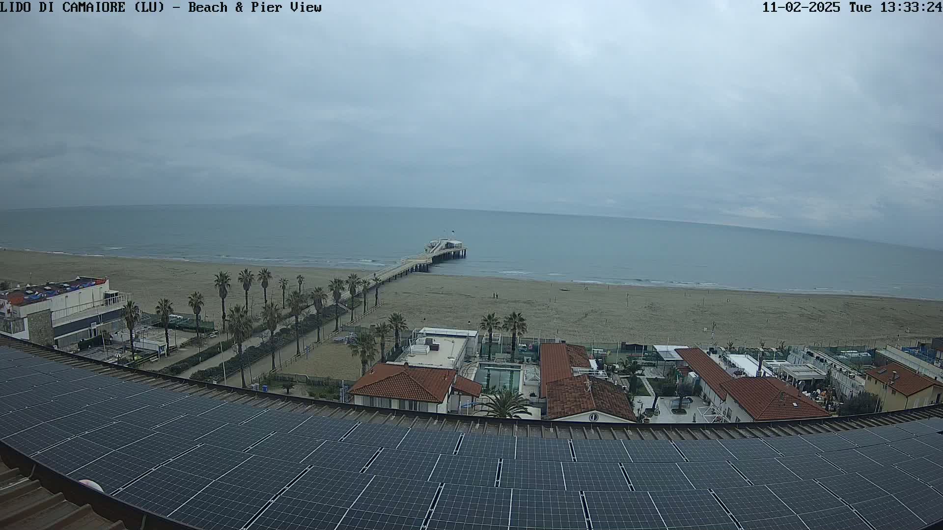 A beach with a pier and several buildings is visible from a rooftop covered in solar panels under an overcast sky.
