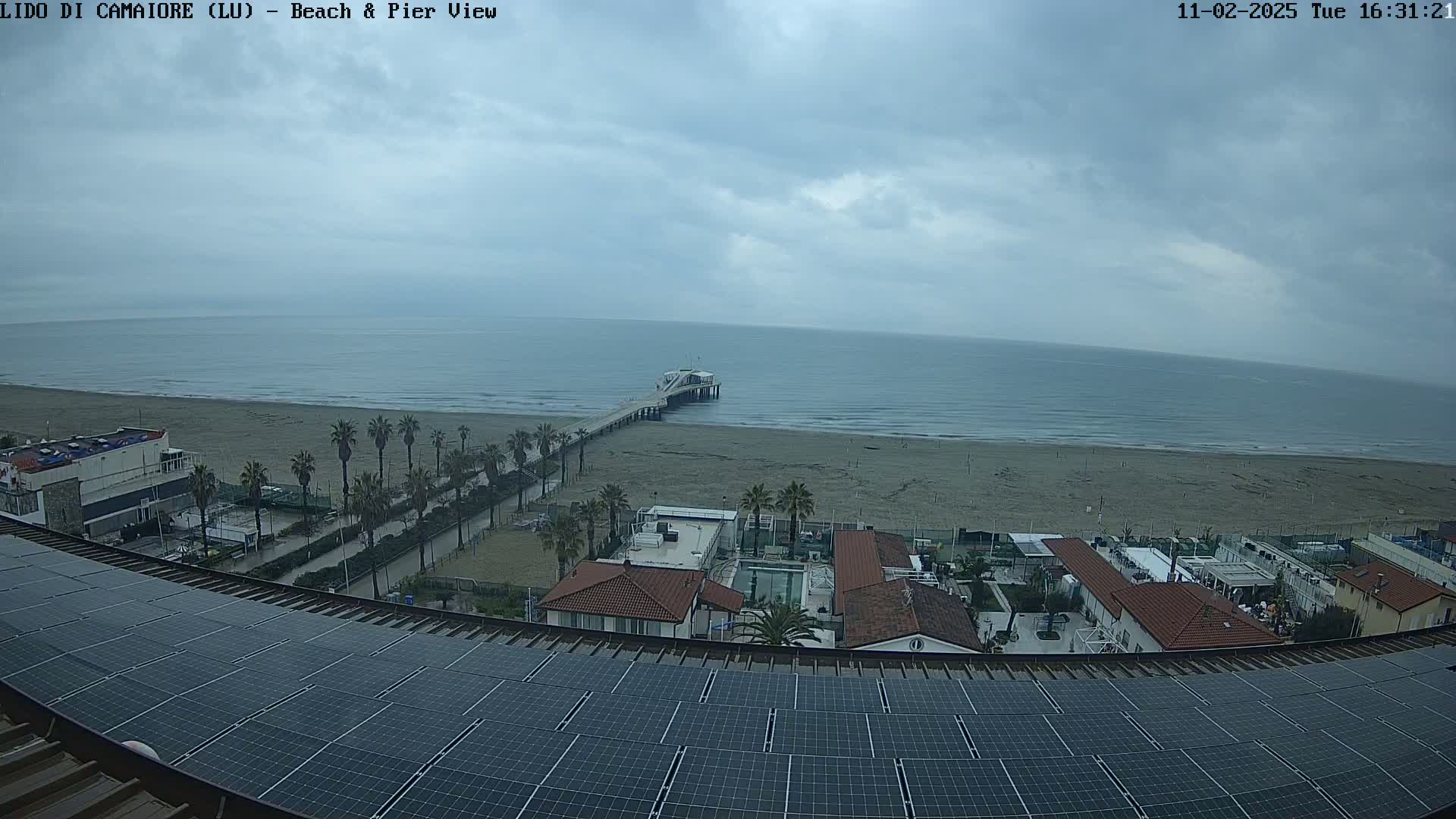 A beach with a pier stretches out to a calm, grey ocean under an overcast sky, viewed from above a building with solar panels on its roof.