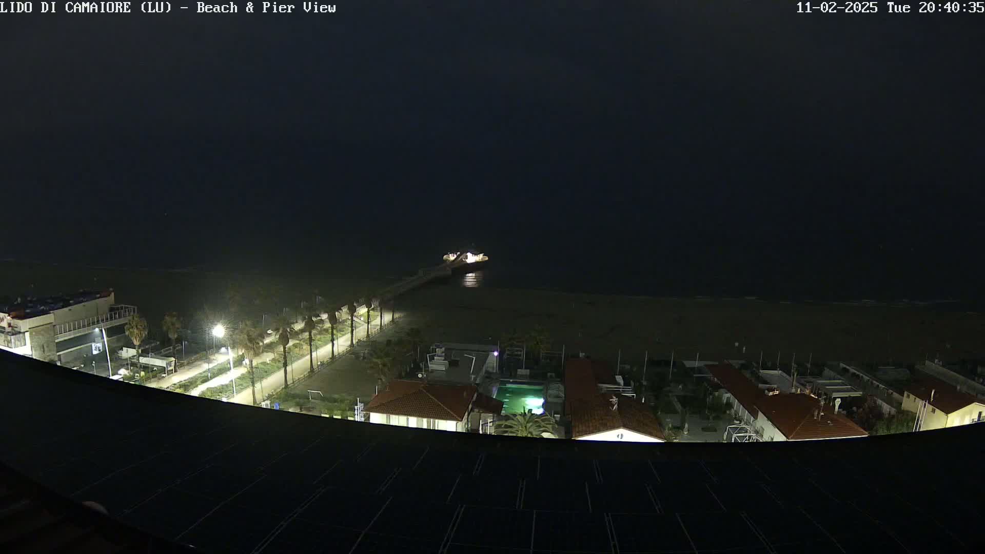 A nighttime view of a beach, pier, and coastal buildings under a dark sky.