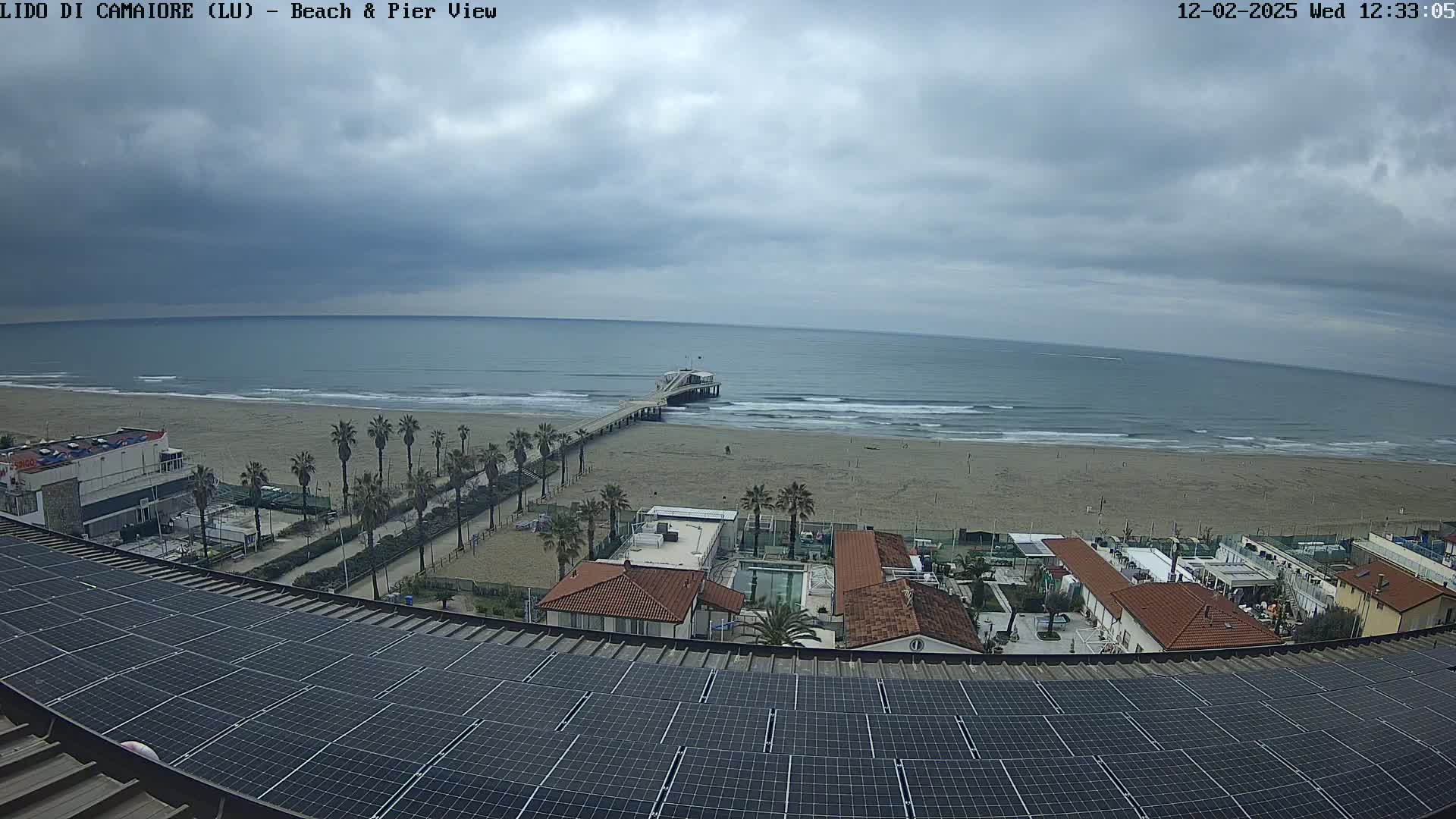A beach with a pier extends into a calm sea under an overcast sky, viewed from a rooftop covered in solar panels.