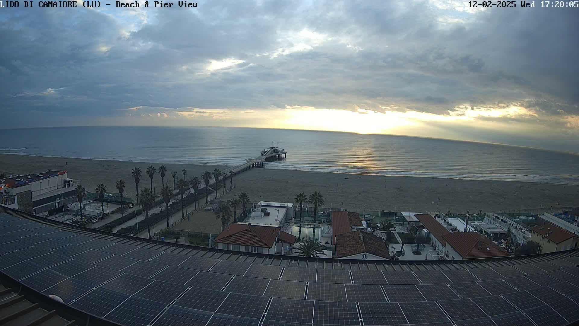 A beach with a pier and buildings is viewed from above under a partly cloudy sky, showing a sunrise over the ocean.