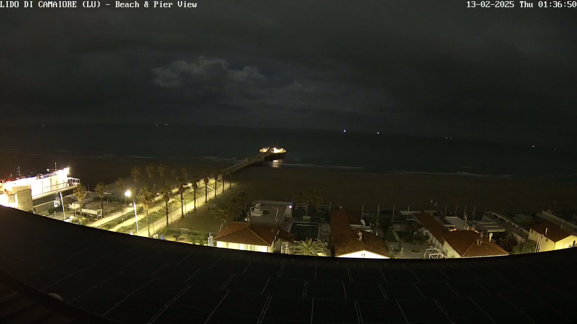A nighttime view of a pier extending into a dark, somewhat cloudy sea, with a beach and buildings illuminated along the shore.