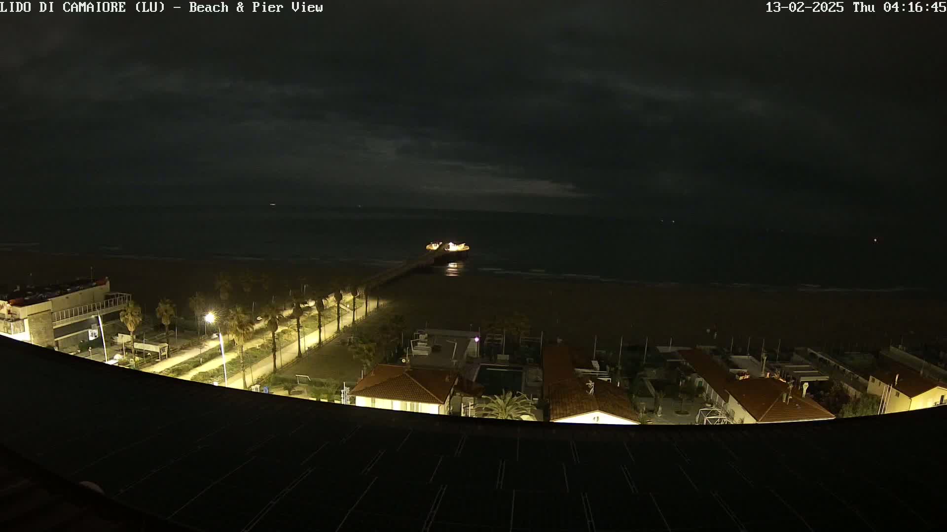 A nighttime view of a beach, pier, and coastal buildings under a dark, cloudy sky.