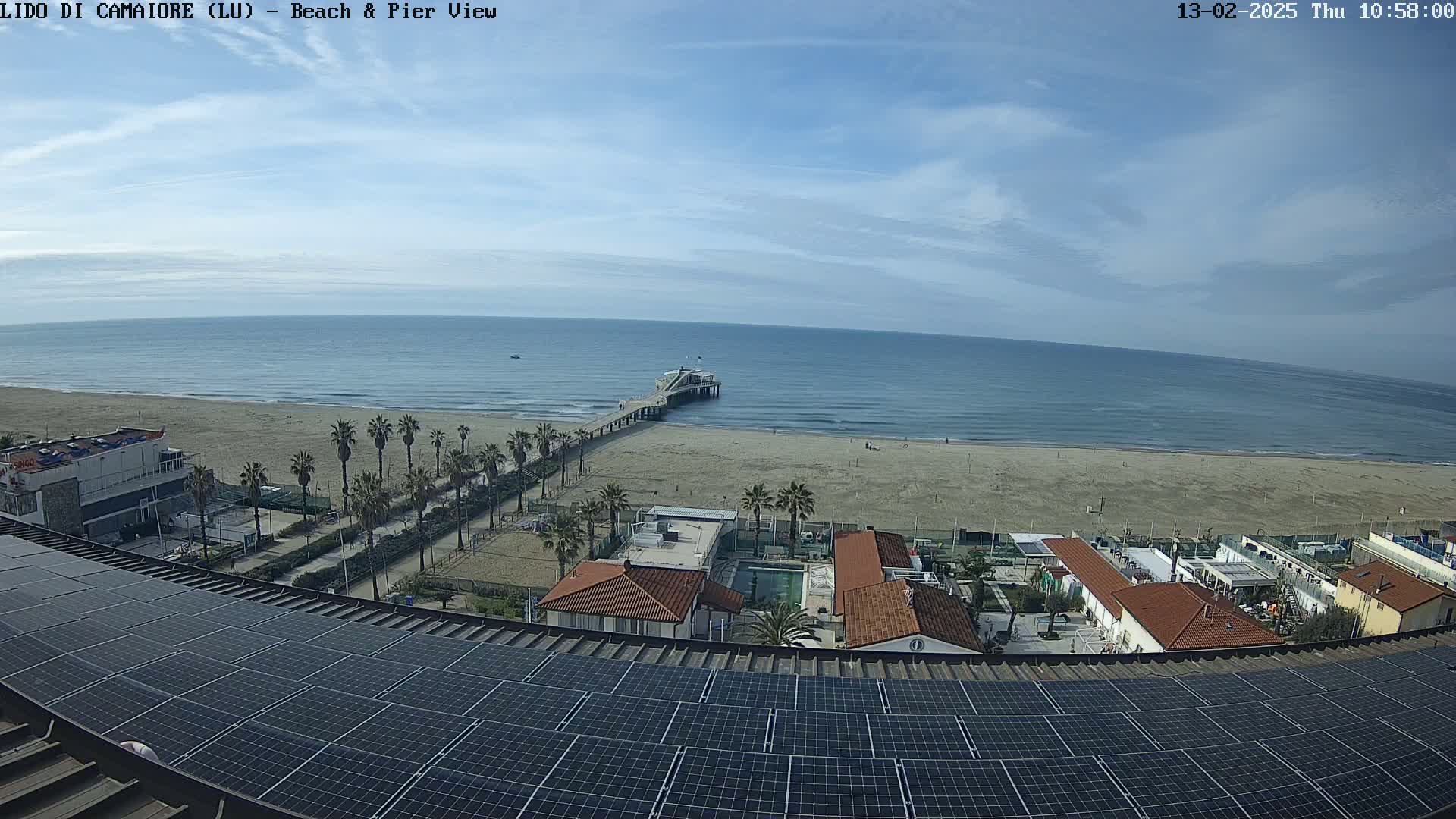 A mostly sunny day shows a beach with a pier, palm trees, buildings, and a large solar panel array on a rooftop overlooking the scene.